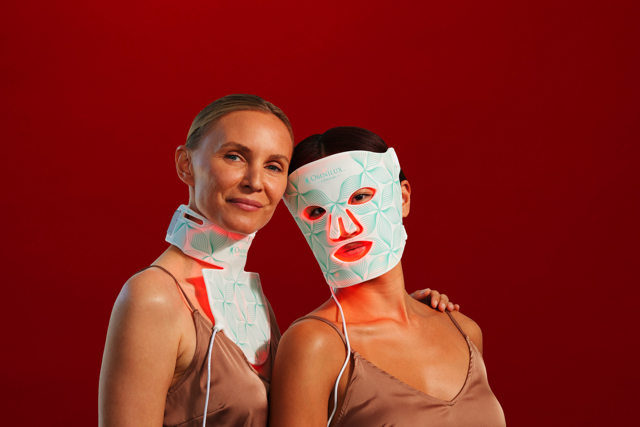 Two women in satin tops, one wearing a white patterned mask, against a red background.