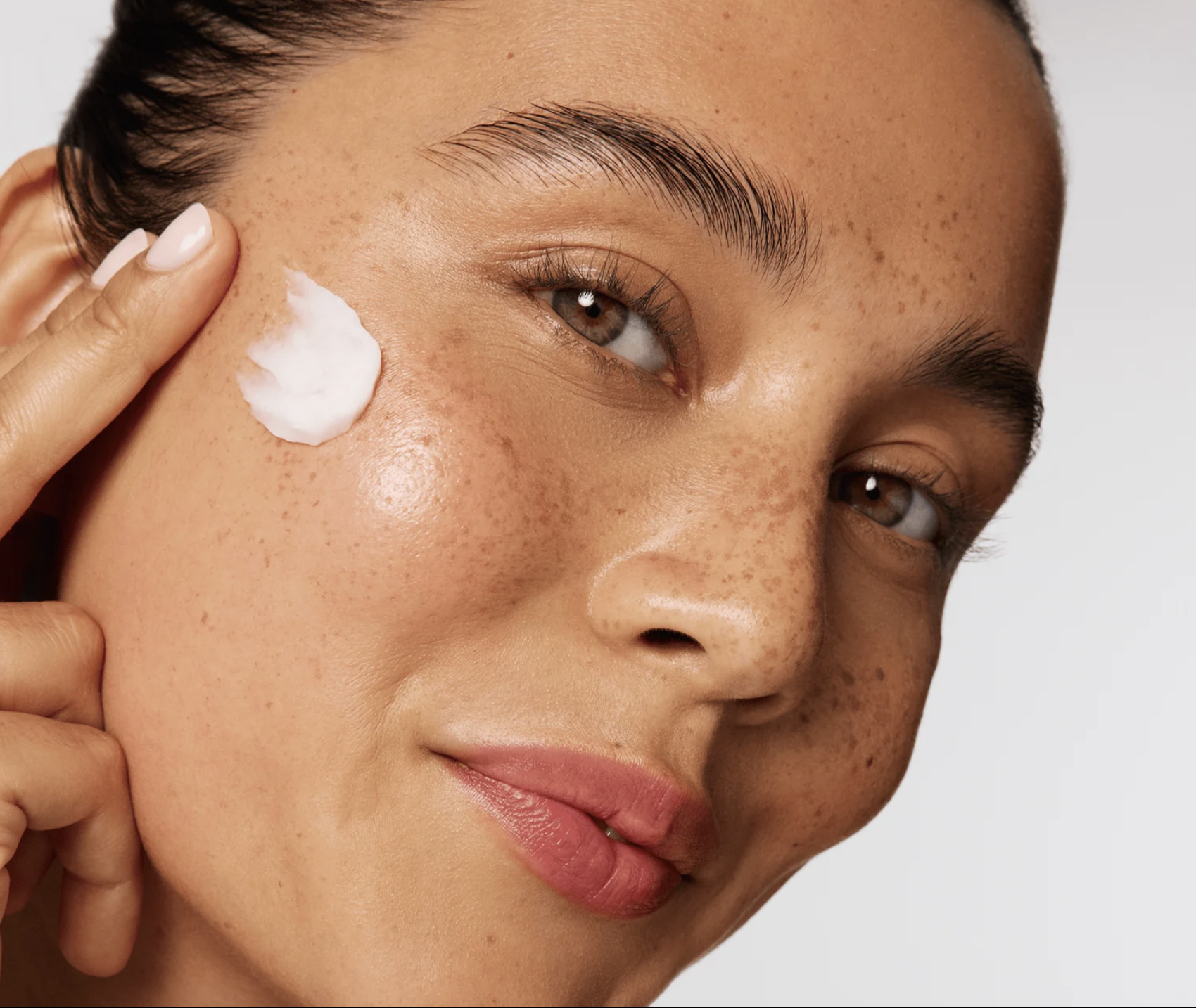 Close-up of a woman with brown eyes applying a dollop of white cream or moisturizer to her cheek.