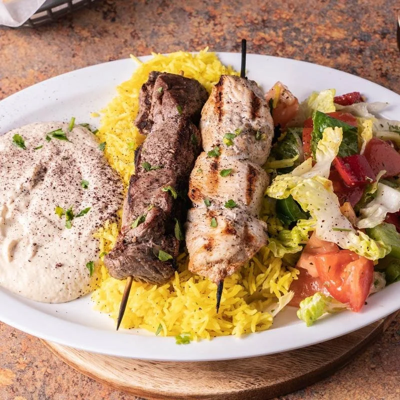 Plate with grilled meat kebabs, yellow rice, salad, and a creamy sauce garnished with herbs.