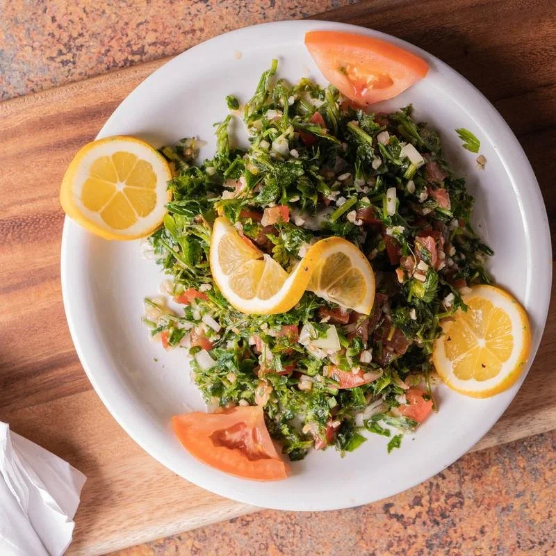 A white plate with a salad containing chopped greens, tomatoes, onions, and lemon slices, on a wooden table.