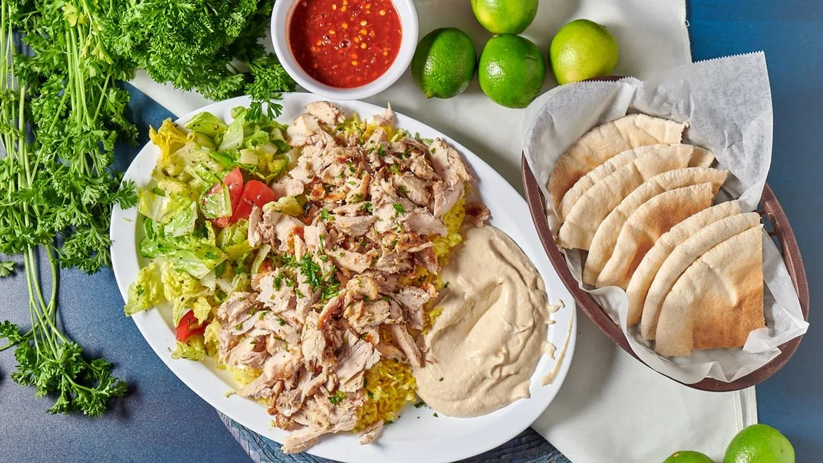 A plate of shredded chicken on rice, salad with lettuce and cherry tomatoes, a side of creamy sauce, and a bowl of red salsa. Surrounding the plate are fresh limes, flatbreads, and bunches of green herbs.
