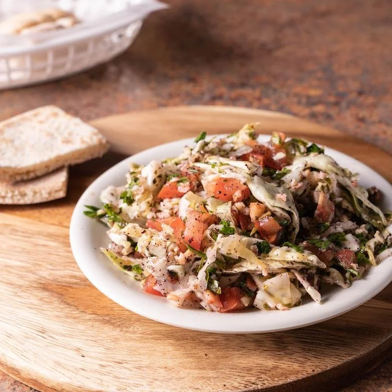 A plate of chopped cabbage salad with tomatoes and herbs on a wooden serving board, with sliced bread in the background.