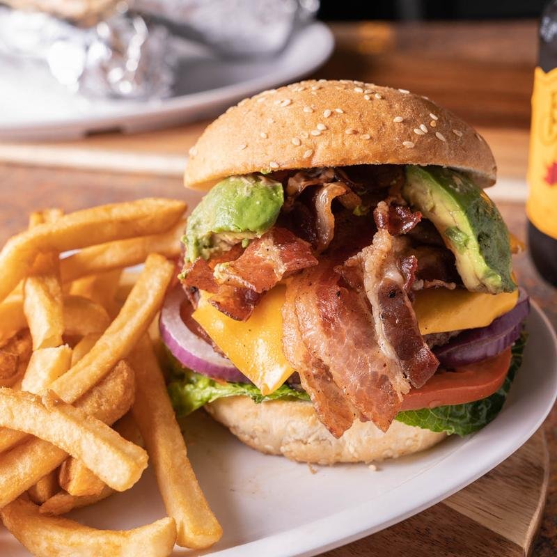 Close-up of a loaded burger with bacon, avocado, cheddar cheese, tomato, onion, lettuce, and a sesame seed bun, served with French fries.