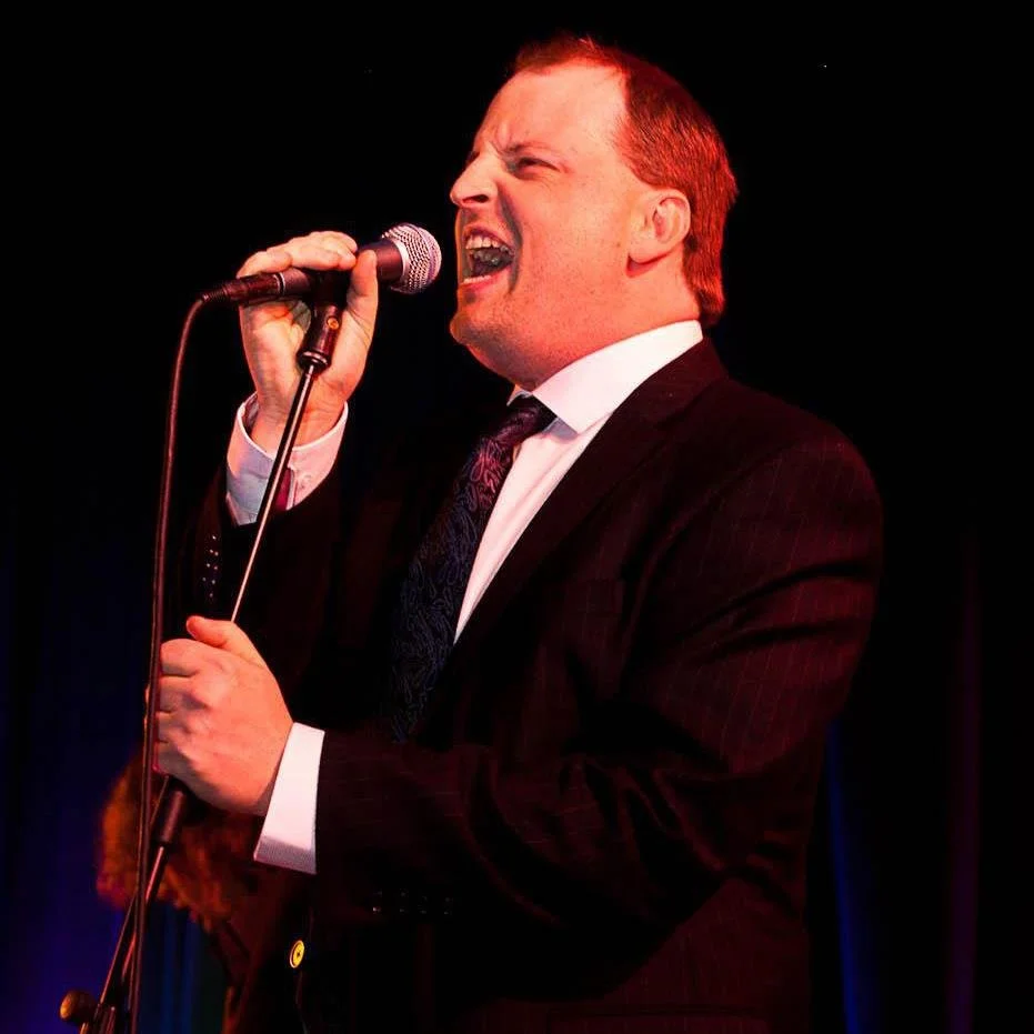 Man singing passionately into a microphone on stage, dressed in a black suit with a white shirt and dark tie.