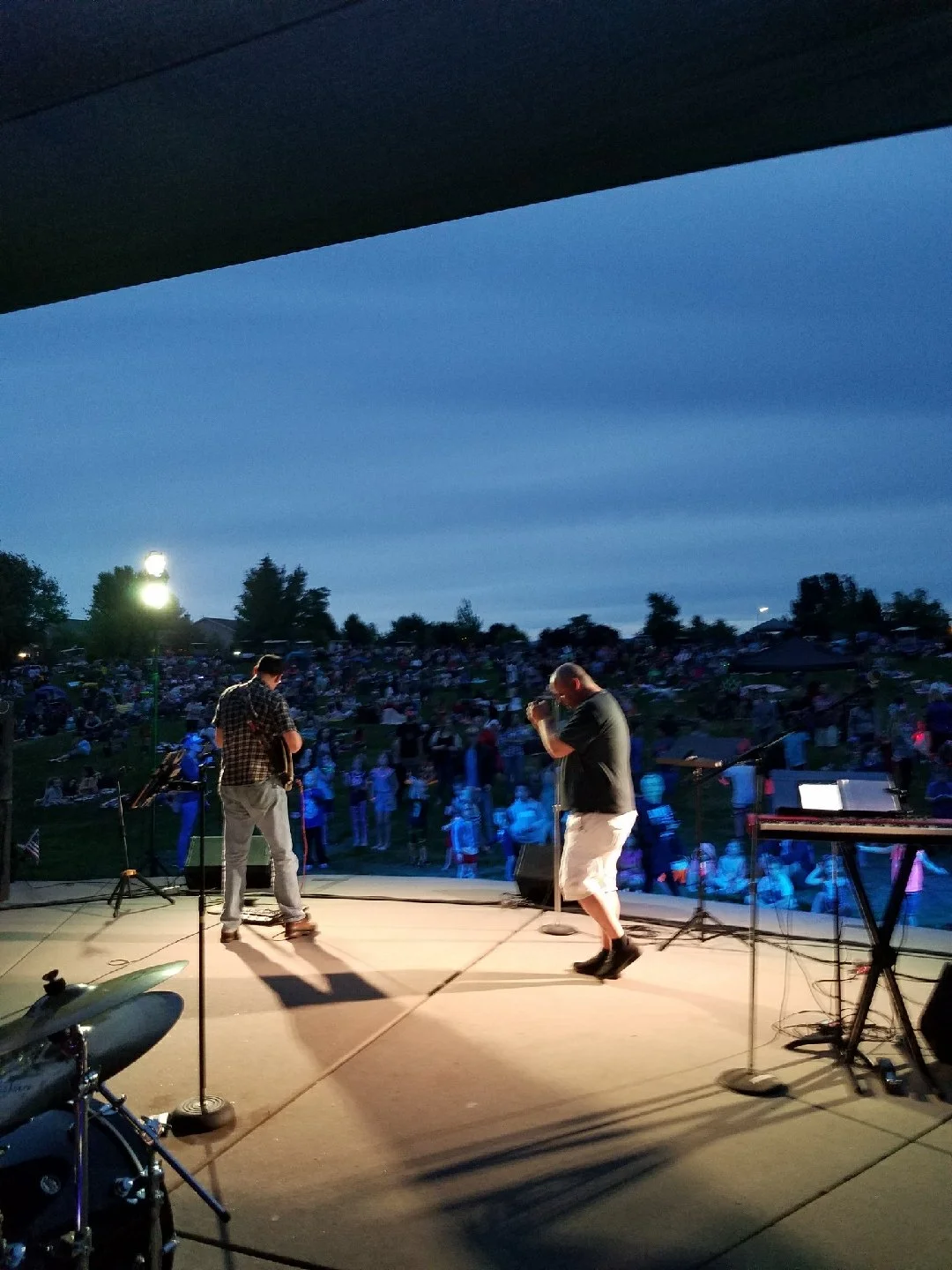 Musicians performing on an outdoor stage at dusk, with a large crowd gathered on a grassy hillside watching the concert.