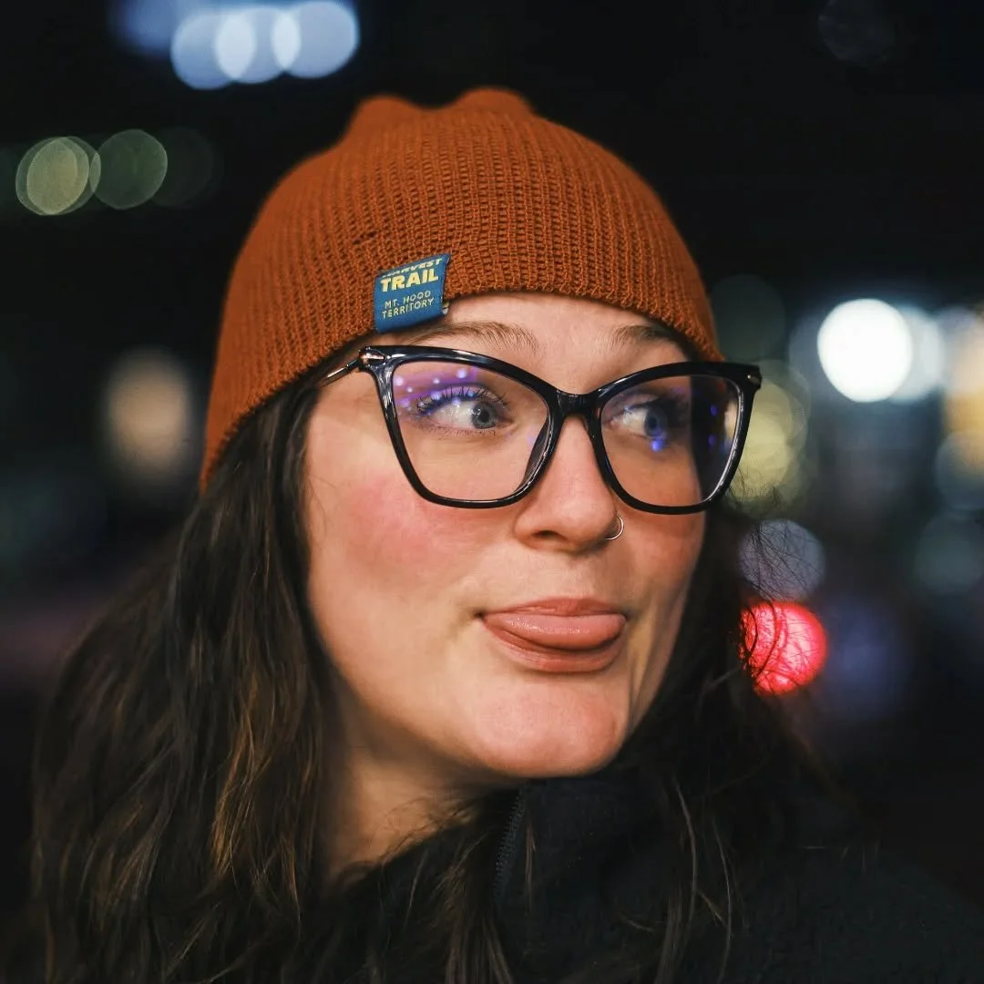A young woman wearing glasses and an orange beanie with a Trail label, making a playful face with her tongue slightly out at night.