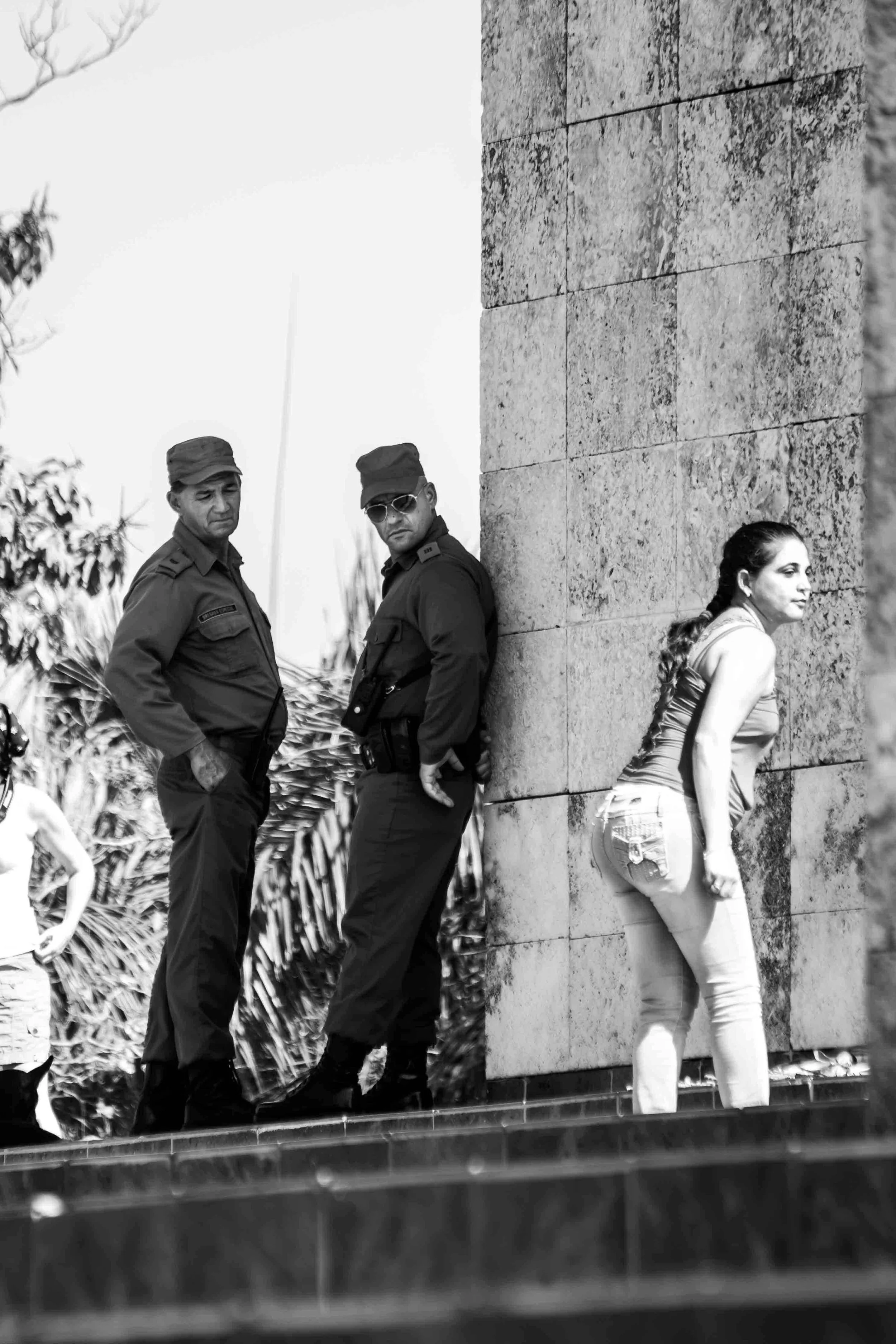 Deux policiers en uniformes debout contre un mur en pierre, une femme en chemisier blanc est assise à côté, toutes les personnes étant visibles en noir et blanc.