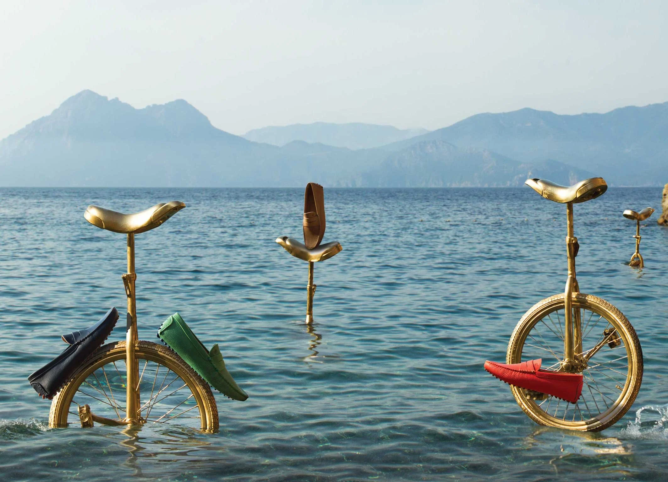 Bicyclette et monocycle roulant sur l'eau, chaussures verte te rouge pédalant, avec des sièges dorés, vue sur la mer et des montagnes en arrière-plan.
