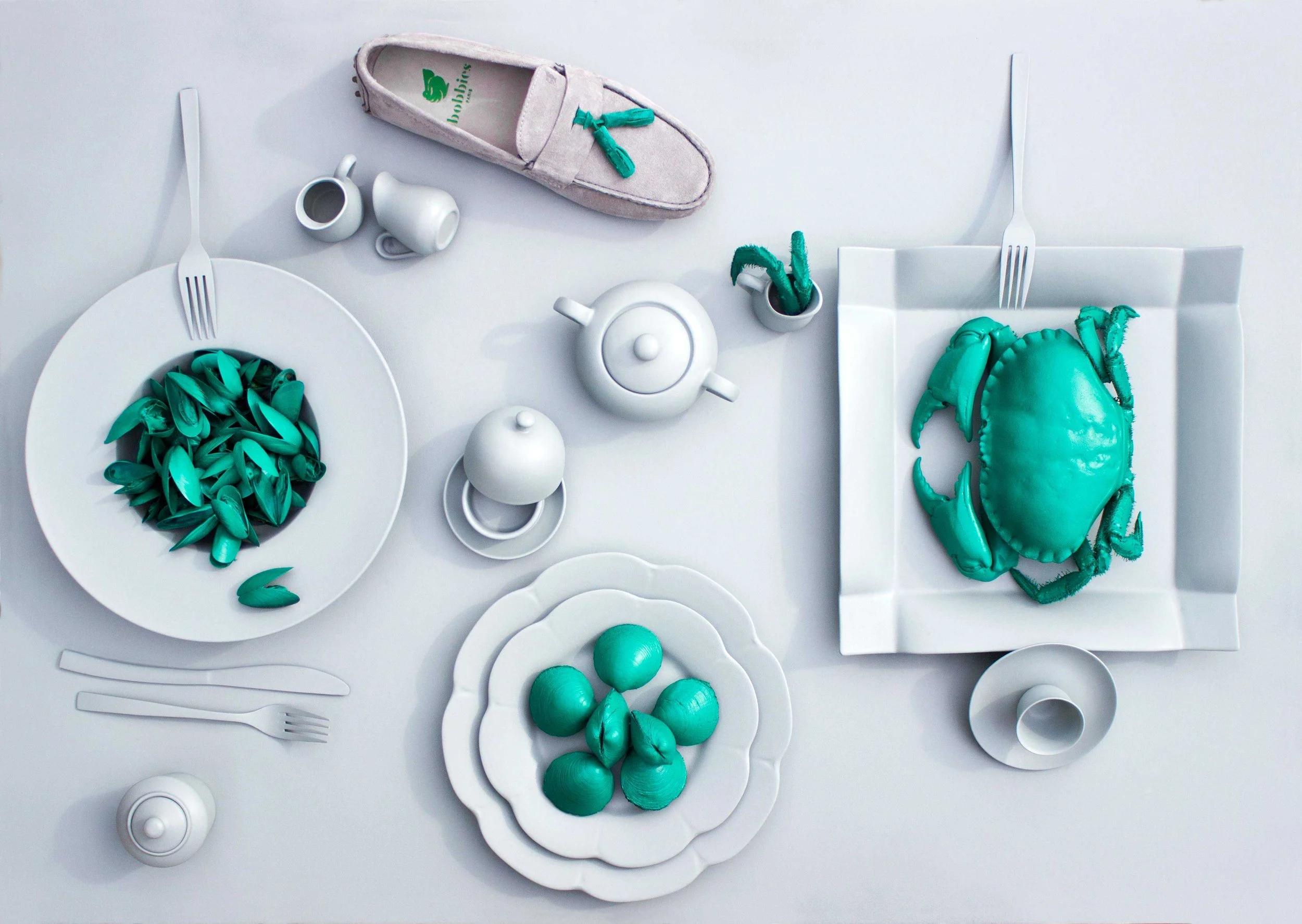 Une table blanche avec des plats, des tasses, une théière et des fourchettes, décorés avec des accessoires de couleur turquoise représentant un crabe et des fruits de mer en plastique, ainsi qu'une chaussure en tissu beige avec un chausson à pompons 
