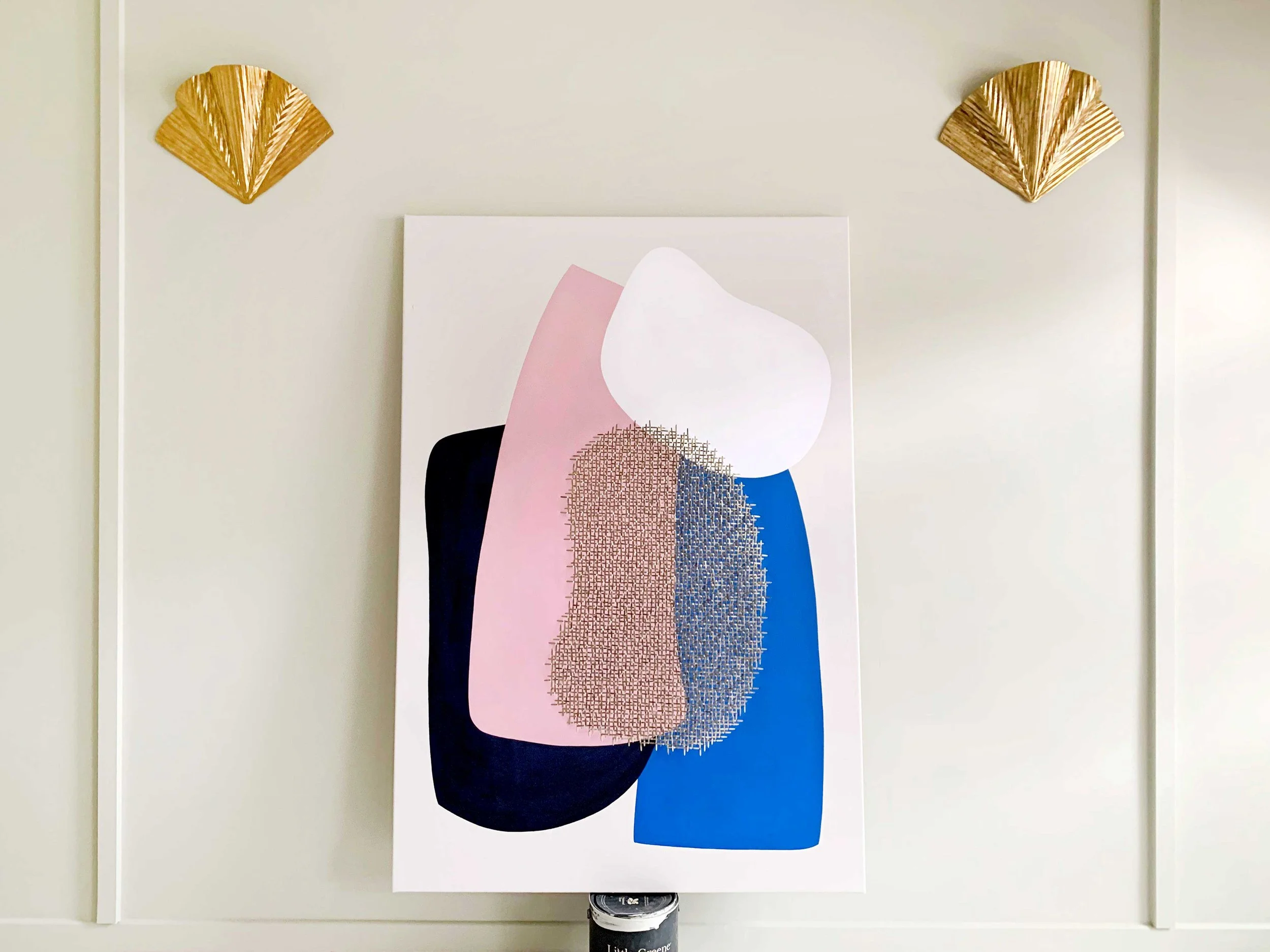Une peinture abstraite avec des formes géométriques en gris, blanc, noir, rose et bleu, appuyée contre un mur blanc. Deux décorations dorées en forme de feuilles ornent le mur au-dessus de la peinture.