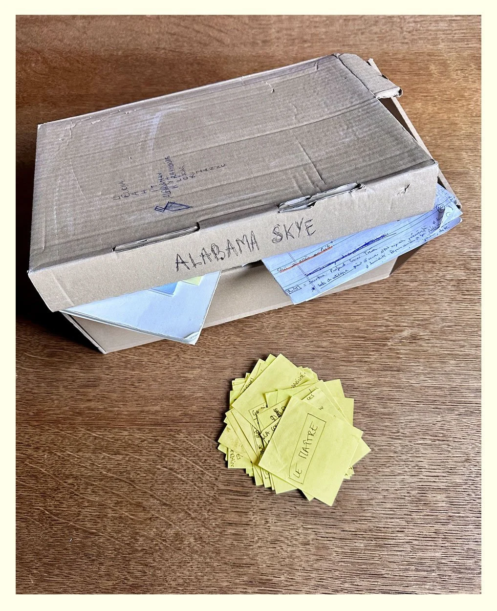 Une boîte en carton contenant des notes manuscrites, avec une petite pile de papiers jaunes et une feuille bleue à côté, sur une table en bois.