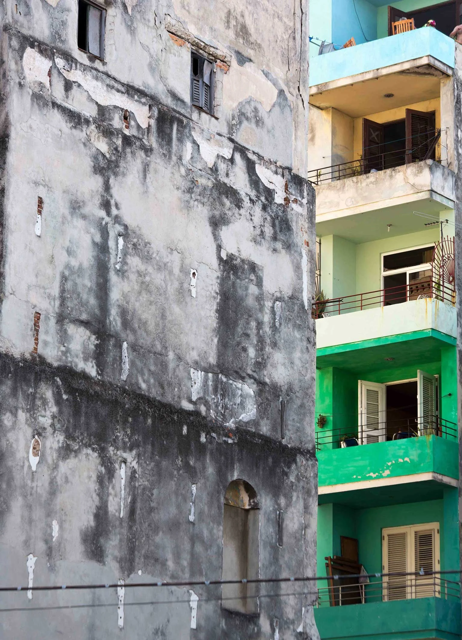 Un bâtiment en mauvais état avec des murs gris dégradés et des fenêtres cassées à gauche, et un bâtiment en bon état avec des balcons verts et des fenêtres ouvertes à droite. technicolor à Cuba