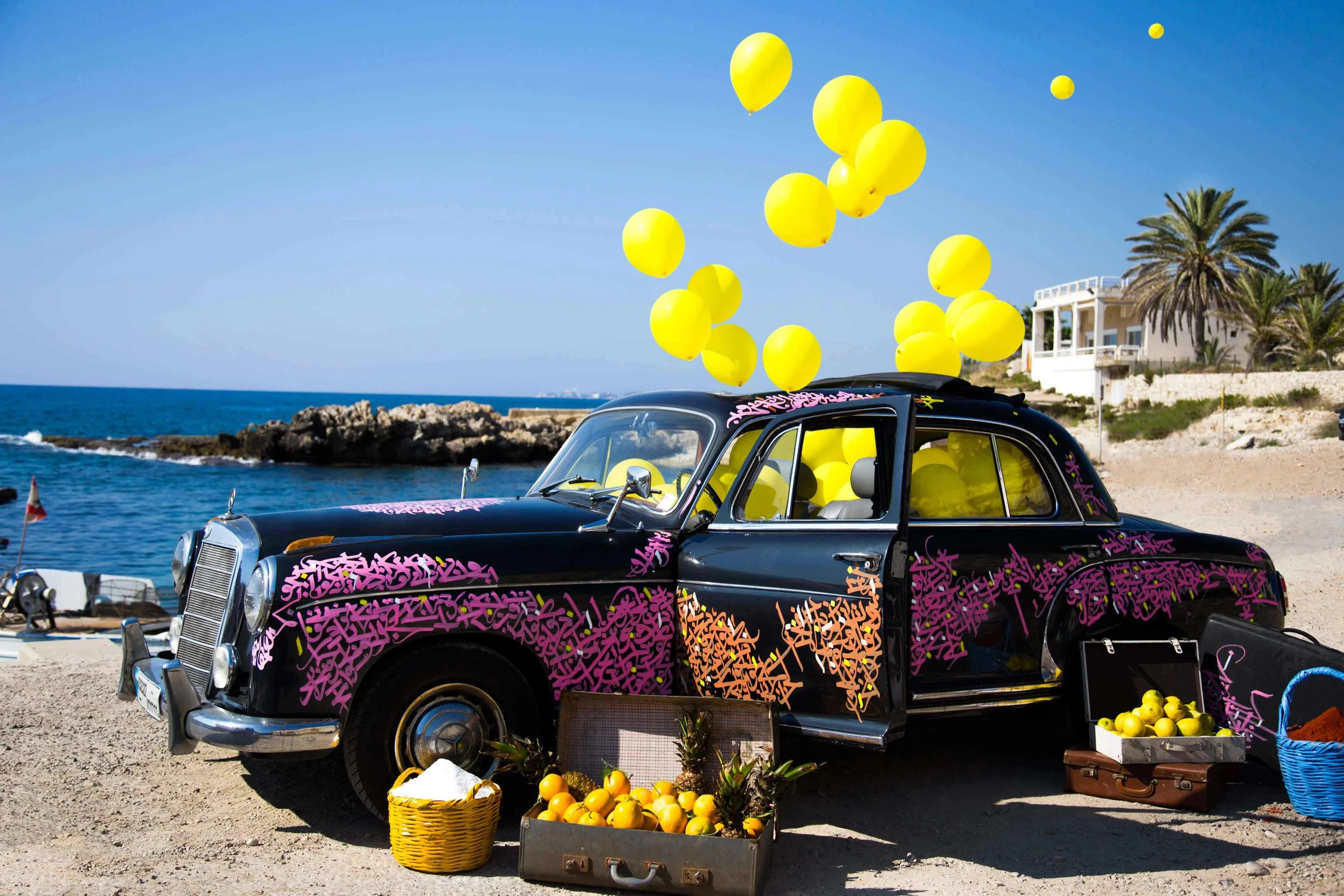 Voiture ancienne noire décorée de calligraphie arabe, avec des ballons jaunes flottant dans les airs et des paniers de fruits jaunes en dessous, avec une maison et des palmiers en arrière-plan