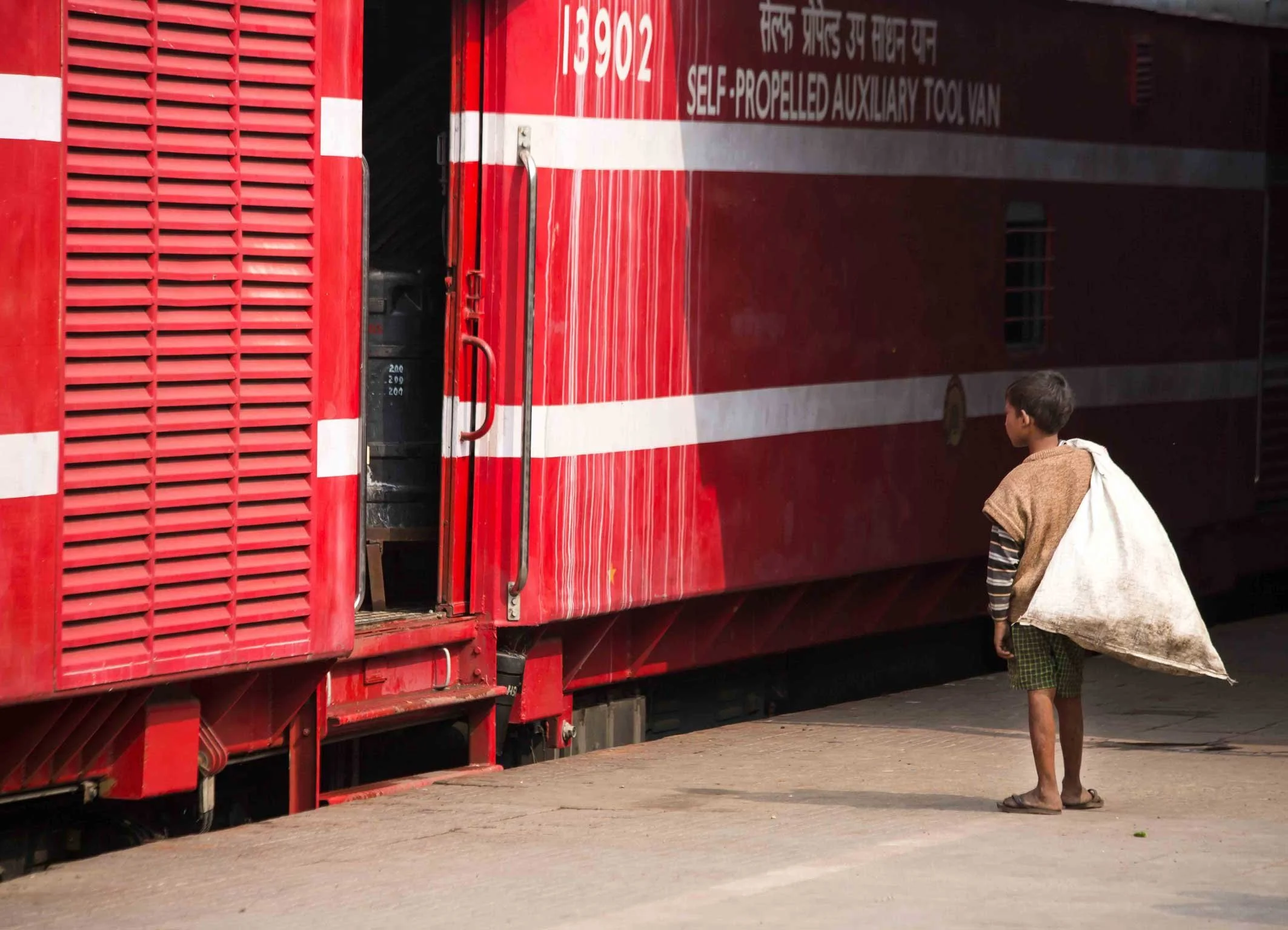 Un garçon indien portant un sac blanc, vu de dos, se tient devant un train rouge avec des bandes blanches.