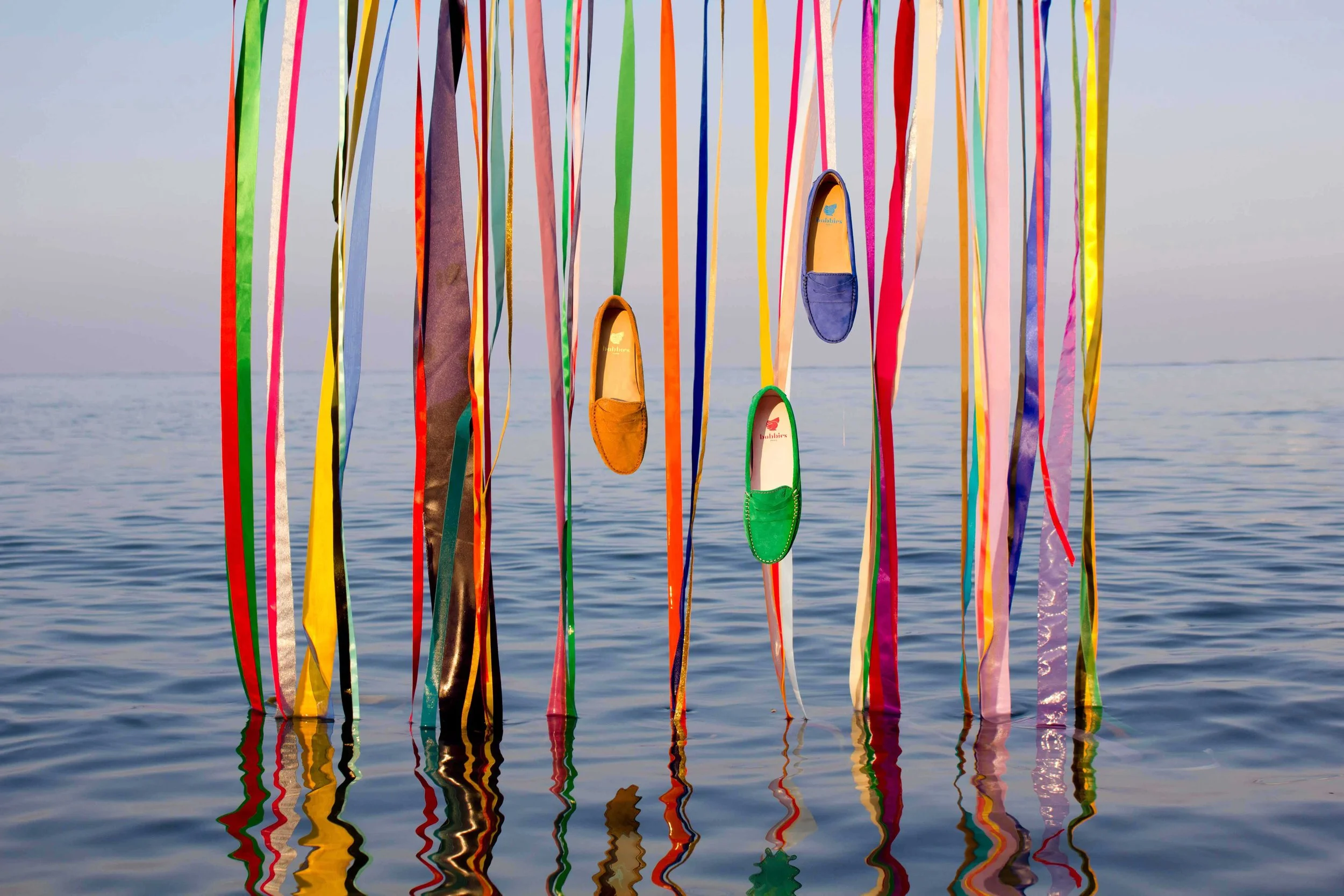 Des chaussures colorées suspendues à des rubans multicolores flottant sur l'eau, créant une installation artistique au bord de la mer.