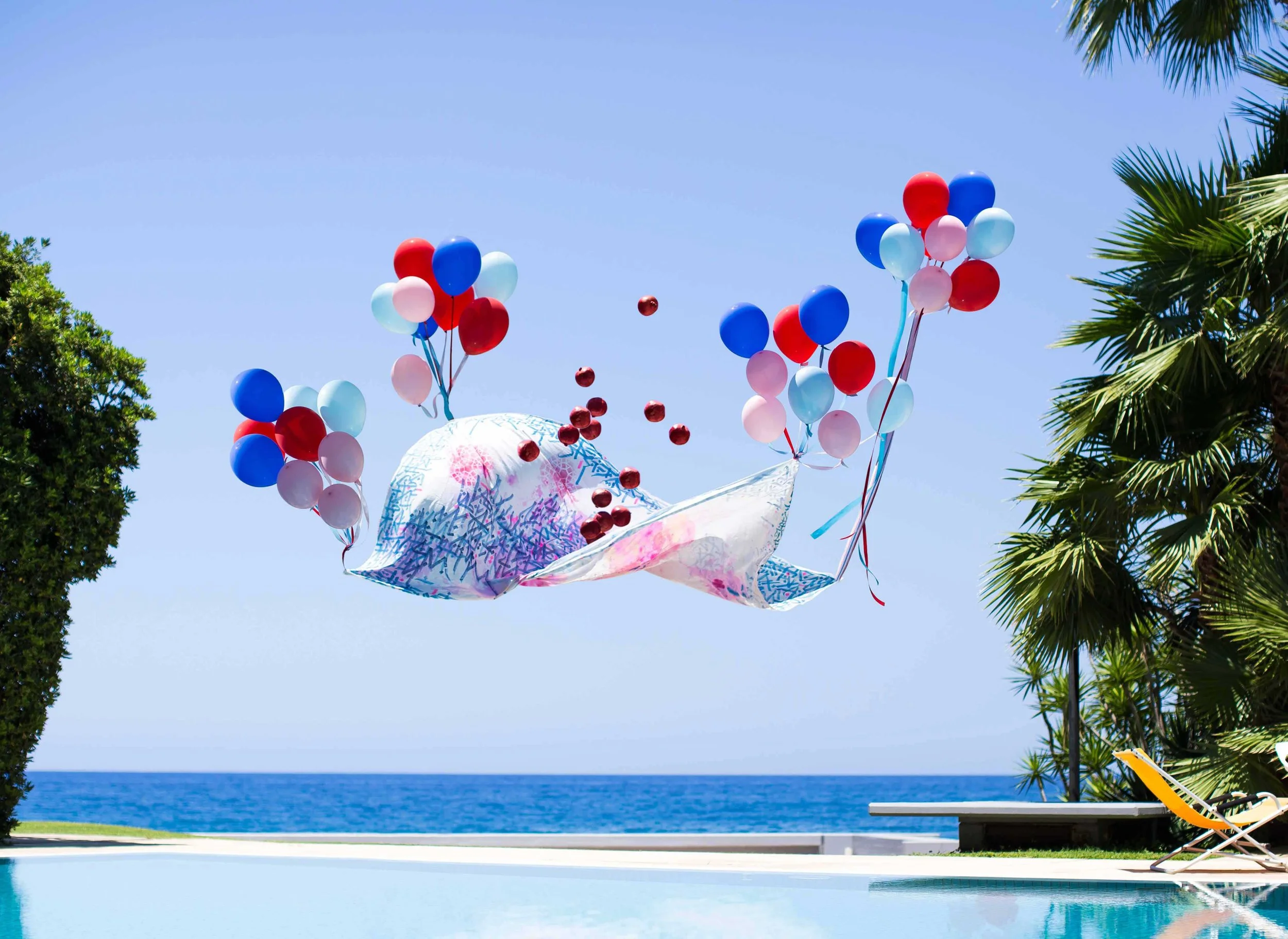 Ballons colorés blanc, rouge et bleu, flottant dans un ciel clair au-dessus d'une plage avec des palmiers et une vue sur la mer.