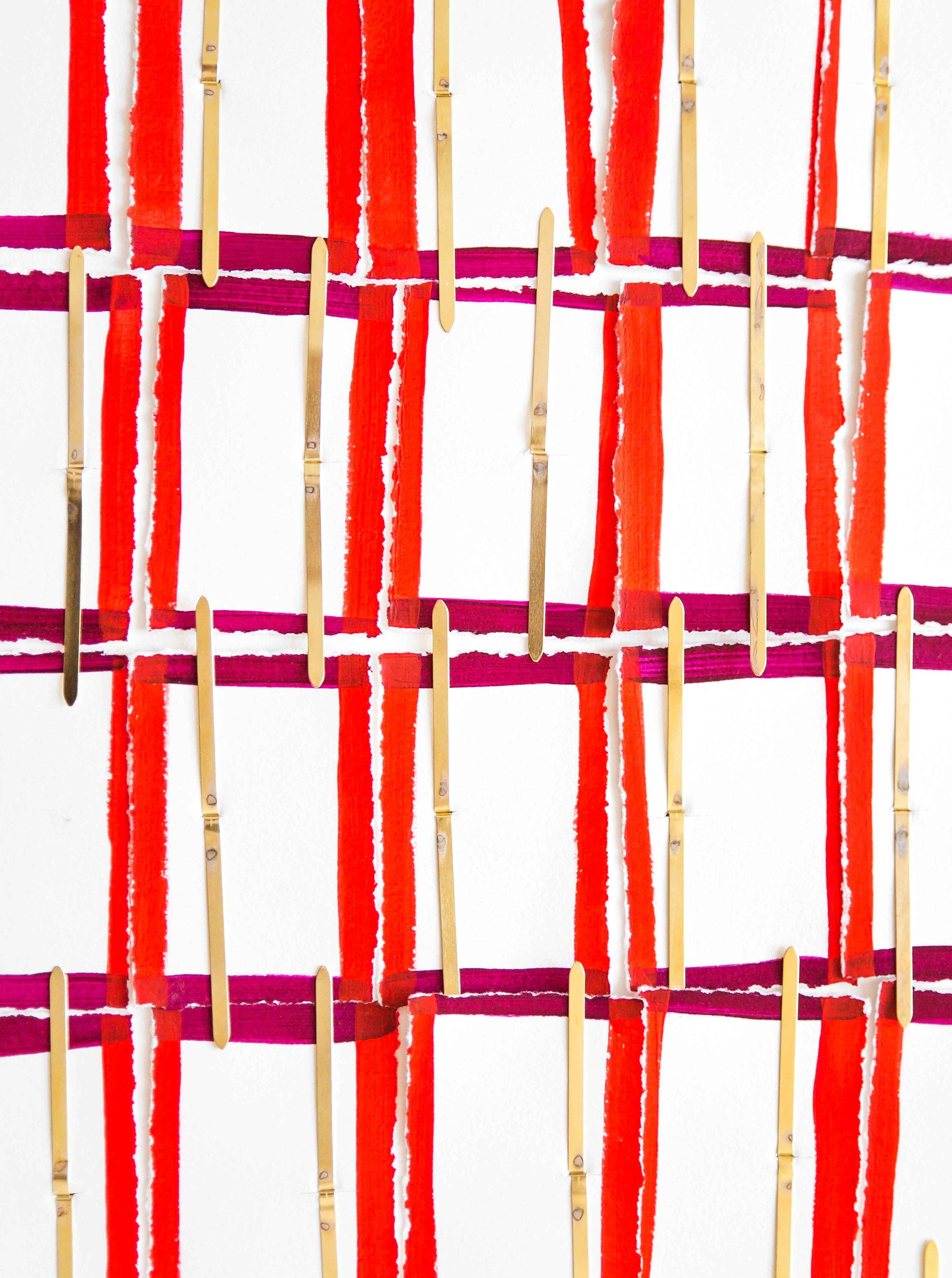Une œuvre d'art abstraite composée de bandes de papier déchiré peintes en rouge, orange et mauve, fixées sur un fond blanc avec des attaches parisiennes.