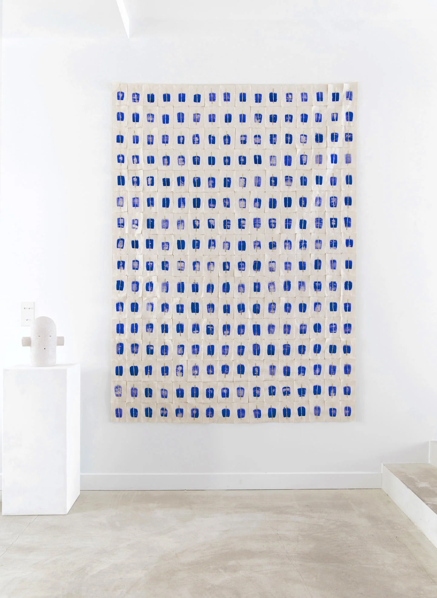 Installation artistique avec une grande œuvre composée de nombreux motifs bleus sur un fond blanc, accompagnée d'une sculpture cylindrique blanche à gauche, exposée à la Galerie Amélie du Chalard.