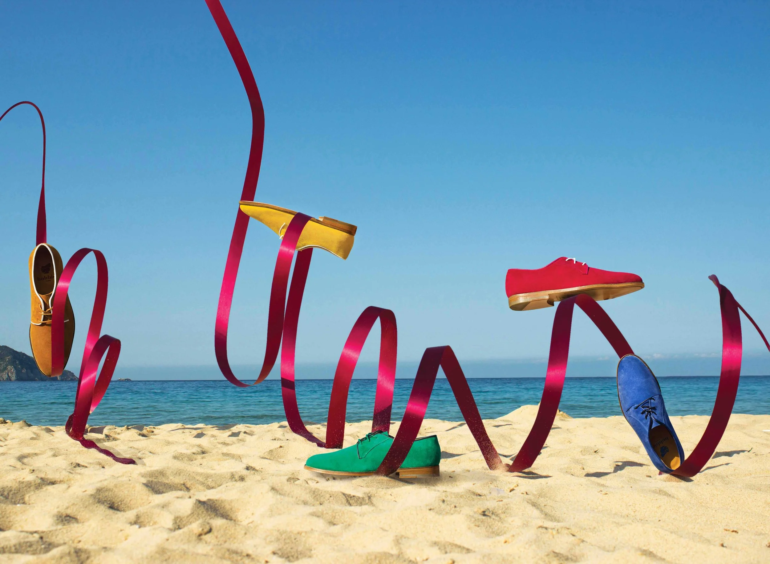 Des chaussures colorées suspendues à des rubans rouges sur une plage avec vue sur la mer et un ciel bleu clair.