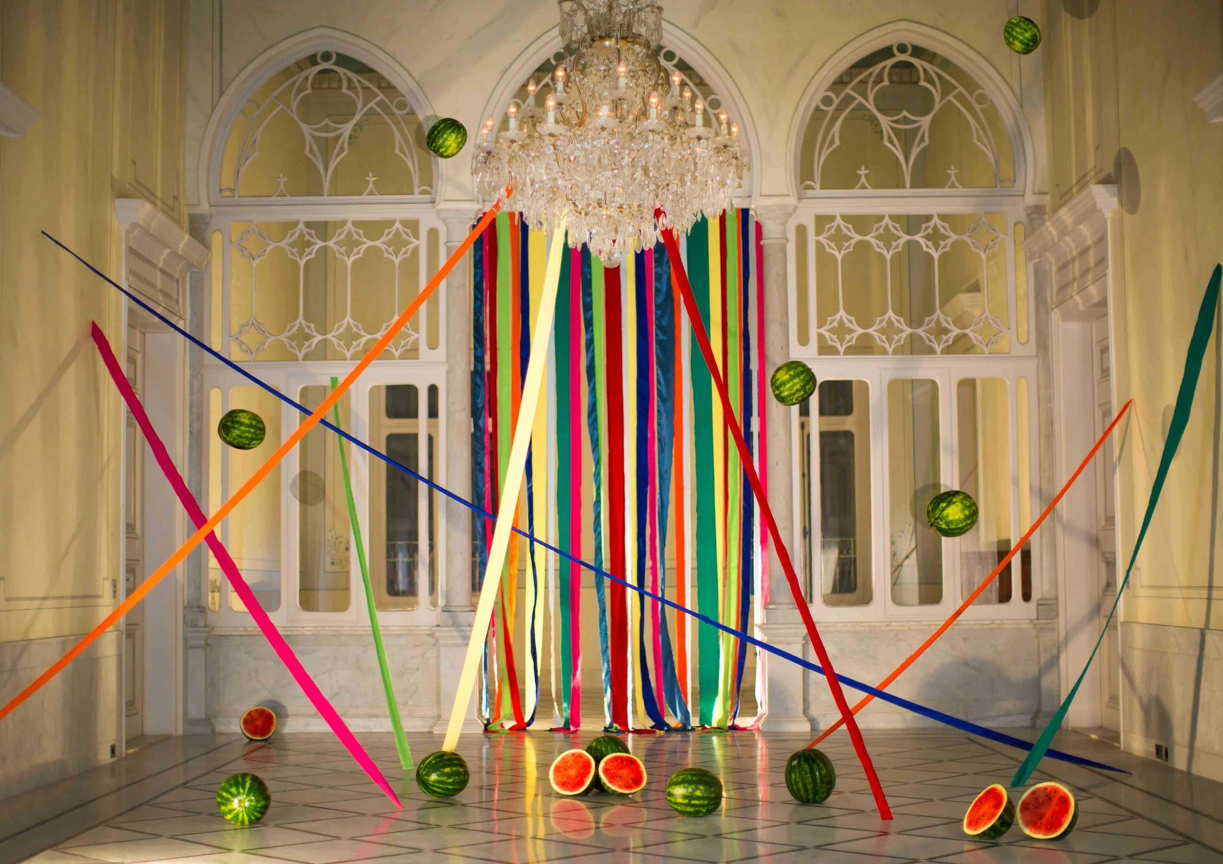 Décor intérieur avec rideau de rubans colorés suspendus, pastèques entières et coupées par terre, et un lustre en cristal suspendu au plafond.