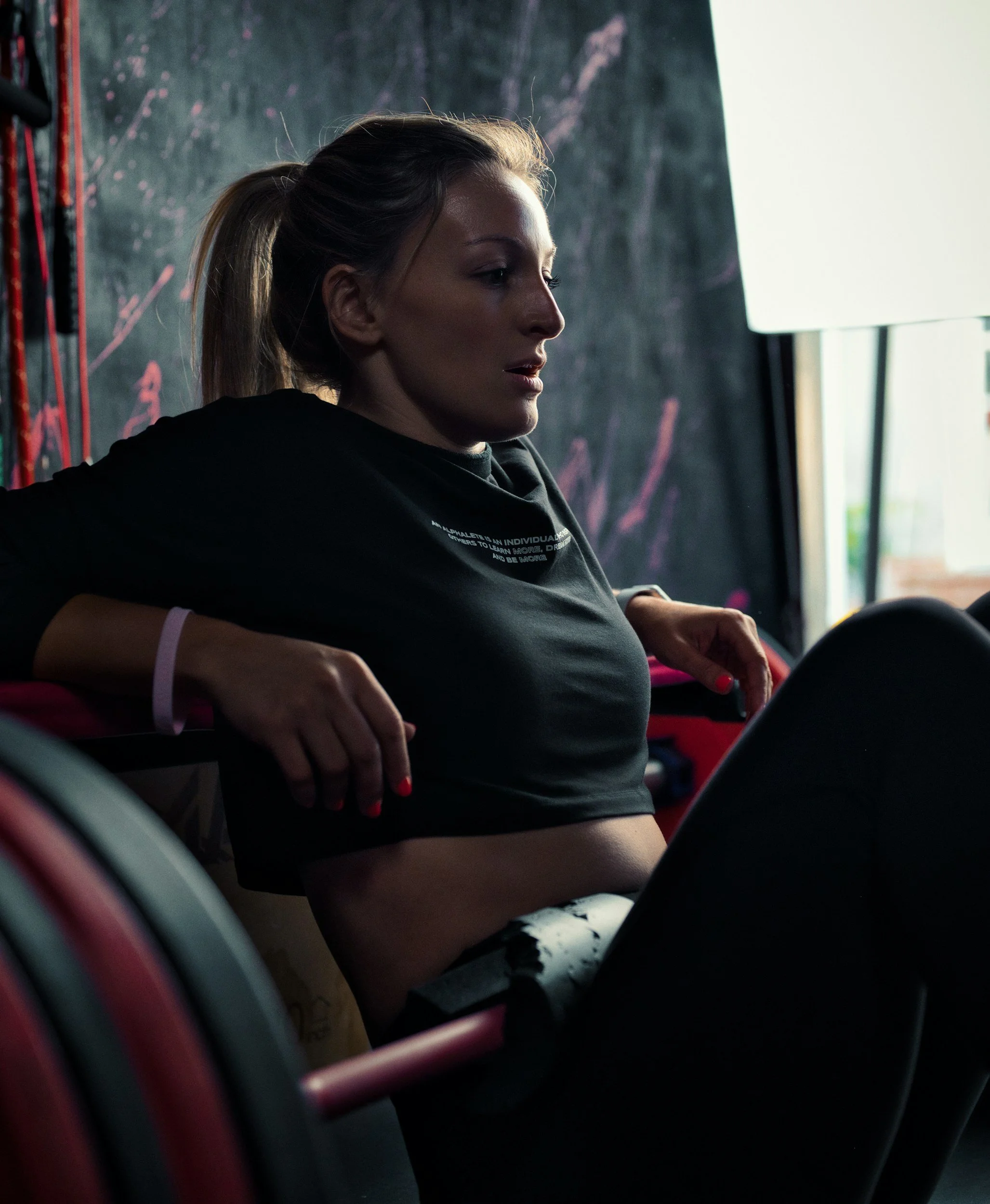 A young woman with blonde hair tied in a ponytail is sitting on a bus or train, looking to her right with a calm expression. She is wearing a black t-shirt with white text and has pink nail polish. The background includes a window and a dark wall with pink markings.