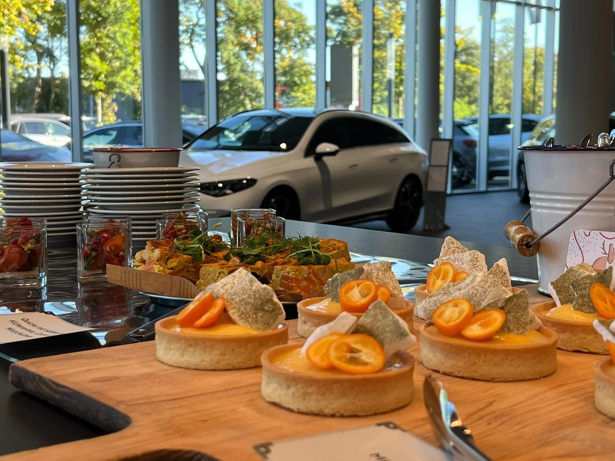 Blick auf eine Theke mit verschiedenen Speisen, darunter belegte Brötchen mit Tomaten, und mehrere leere Teller. Im Hintergrund große Fenster mit Blick auf parkende Autos und Bäume.