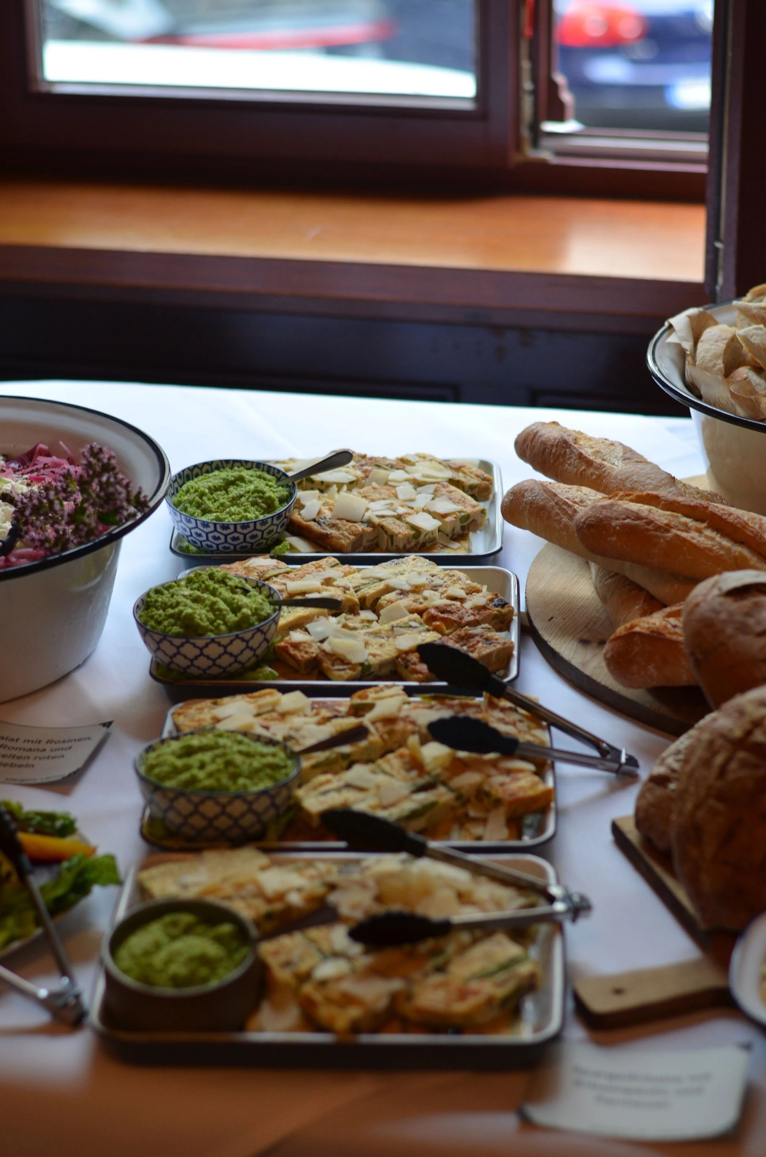 Verschiedene italienische Speisen auf einem Buffet, darunter frisch gebackene Baguettes, Pizzastücke mit Käse, grüne Creme in kleinen Schalen, Salat mit Blumen als Dekoration, vor einem Fenster