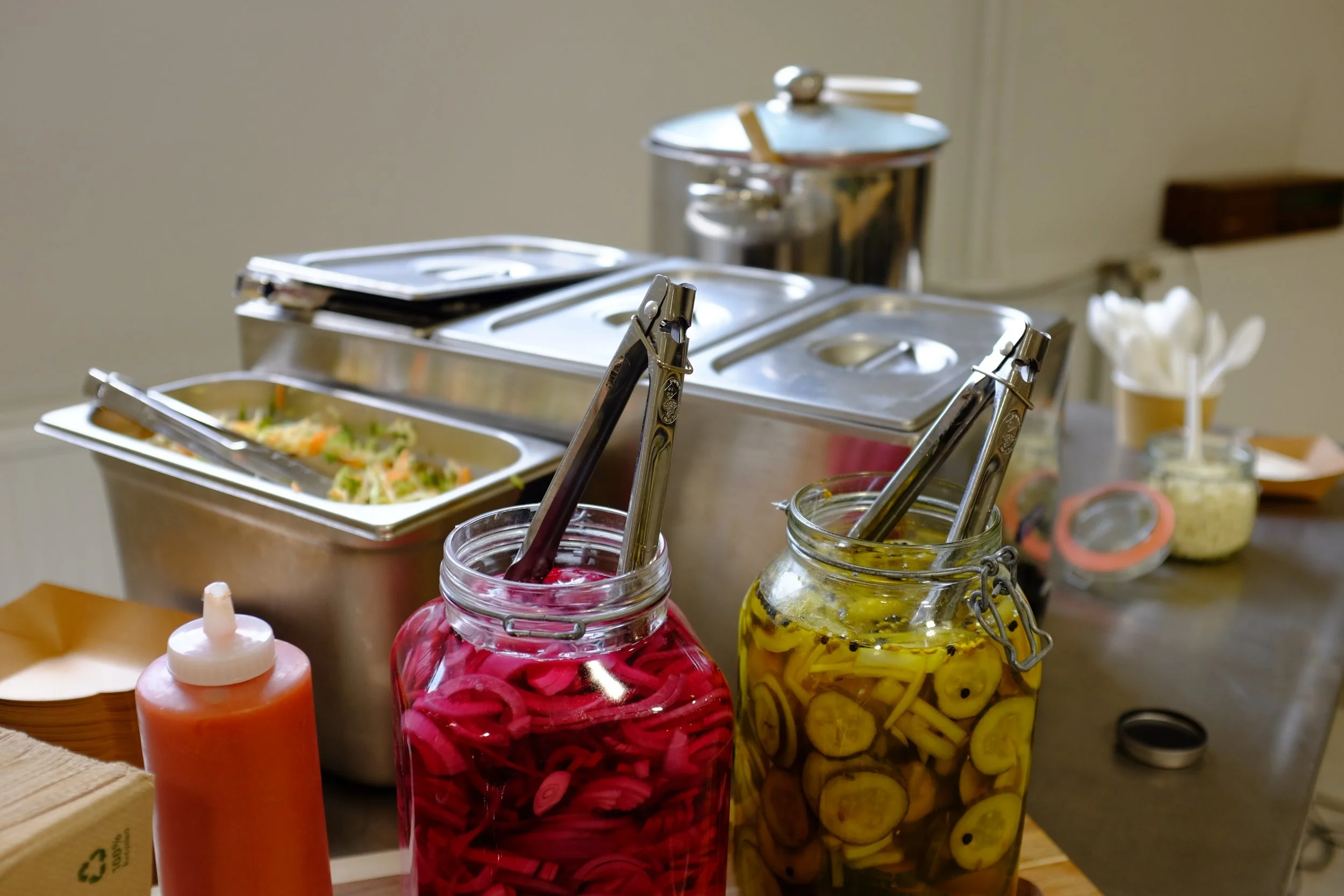 Verschiedene Glasvasen mit eingelegtem Gemüse wie Gurken und roten Zwiebeln, sowie Saucenflaschen auf einem Buffet-Tisch.