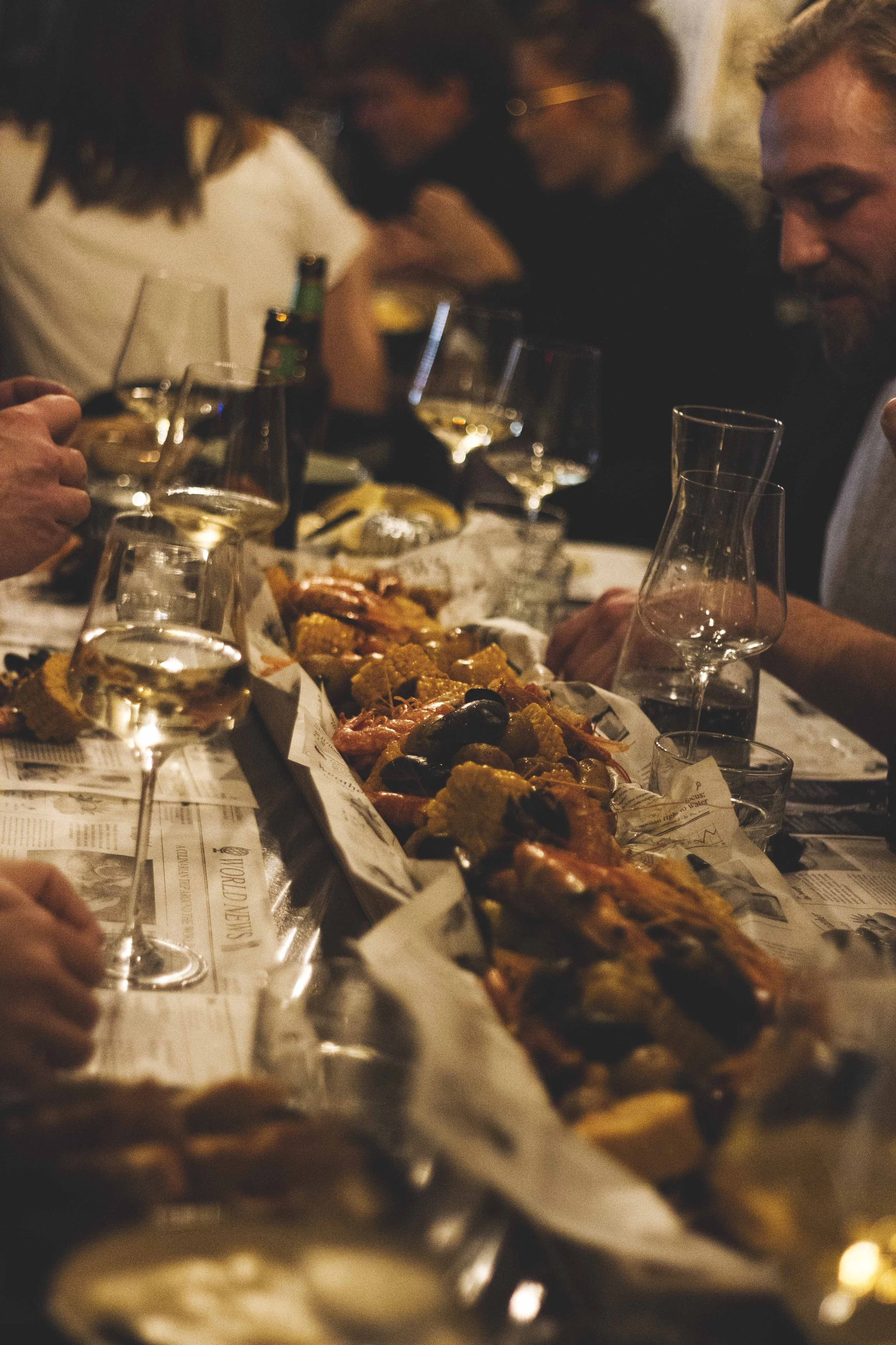 Menschen sitzen bei einem Abendessen mit Wein und Meeresfrüchten, die auf Papier ausgelegt sind, in einer gemütlichen, dunklen Atmosphäre.