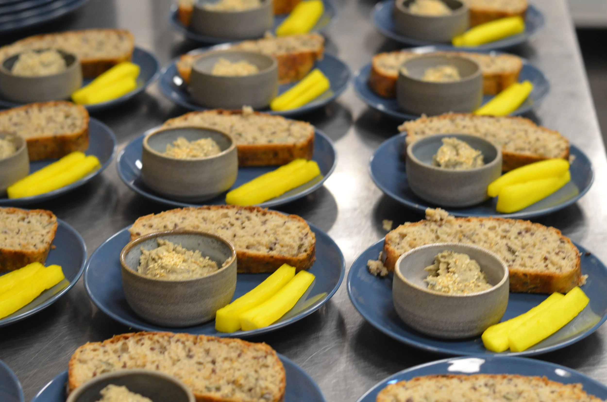 Mehrere blaue Teller mit Schnittbrot, kleinen Schälchen mit Aufstrich, und Gurkenscheiben, auf einer Metalloberfläche.