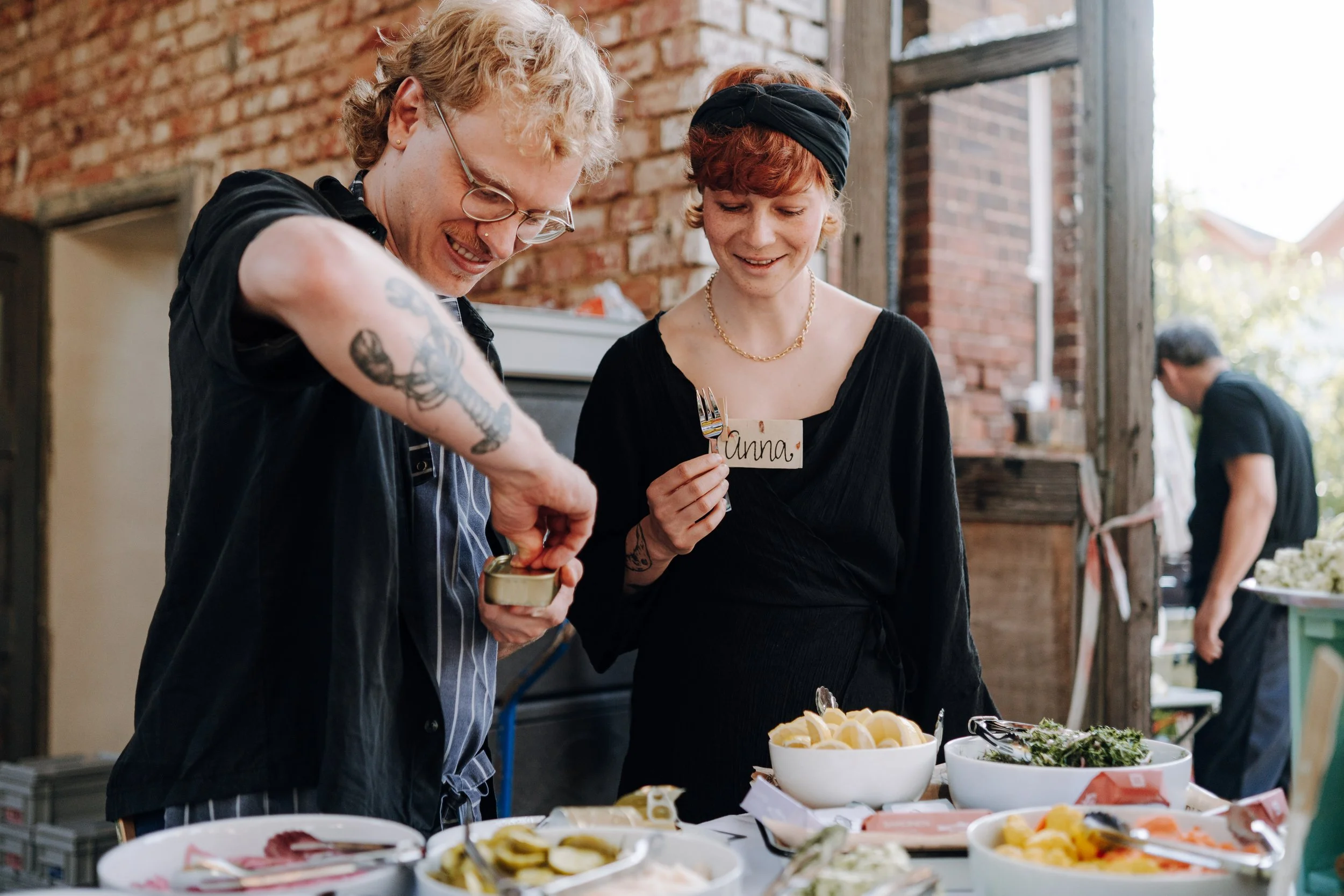 Zwei Menschen, ein Mann mit Blondem, lockigem Haar und Tattoos an den Armen, und eine Frau mit roten Haaren und schwarzem Stirnband, stehen am Buffet und bedienen sich an verschiedenen Speisen.