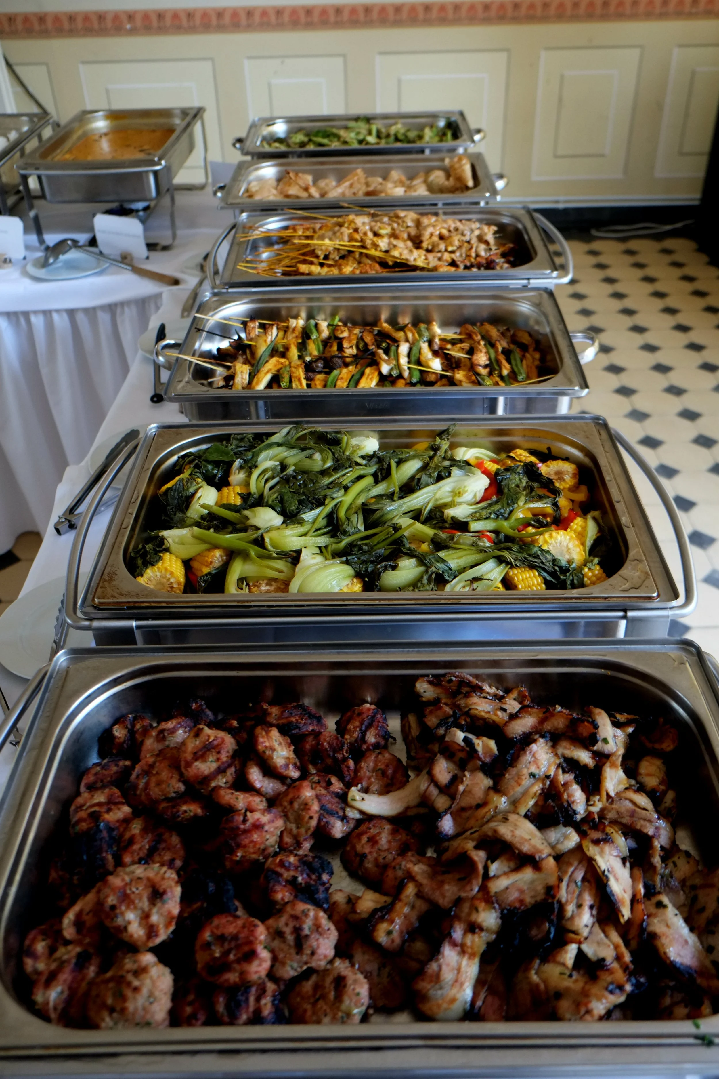 Verschiedene Speisen in Buffet-Containern, darunter gegrilltes Fleisch, Gemüse und Fleischbällchen.