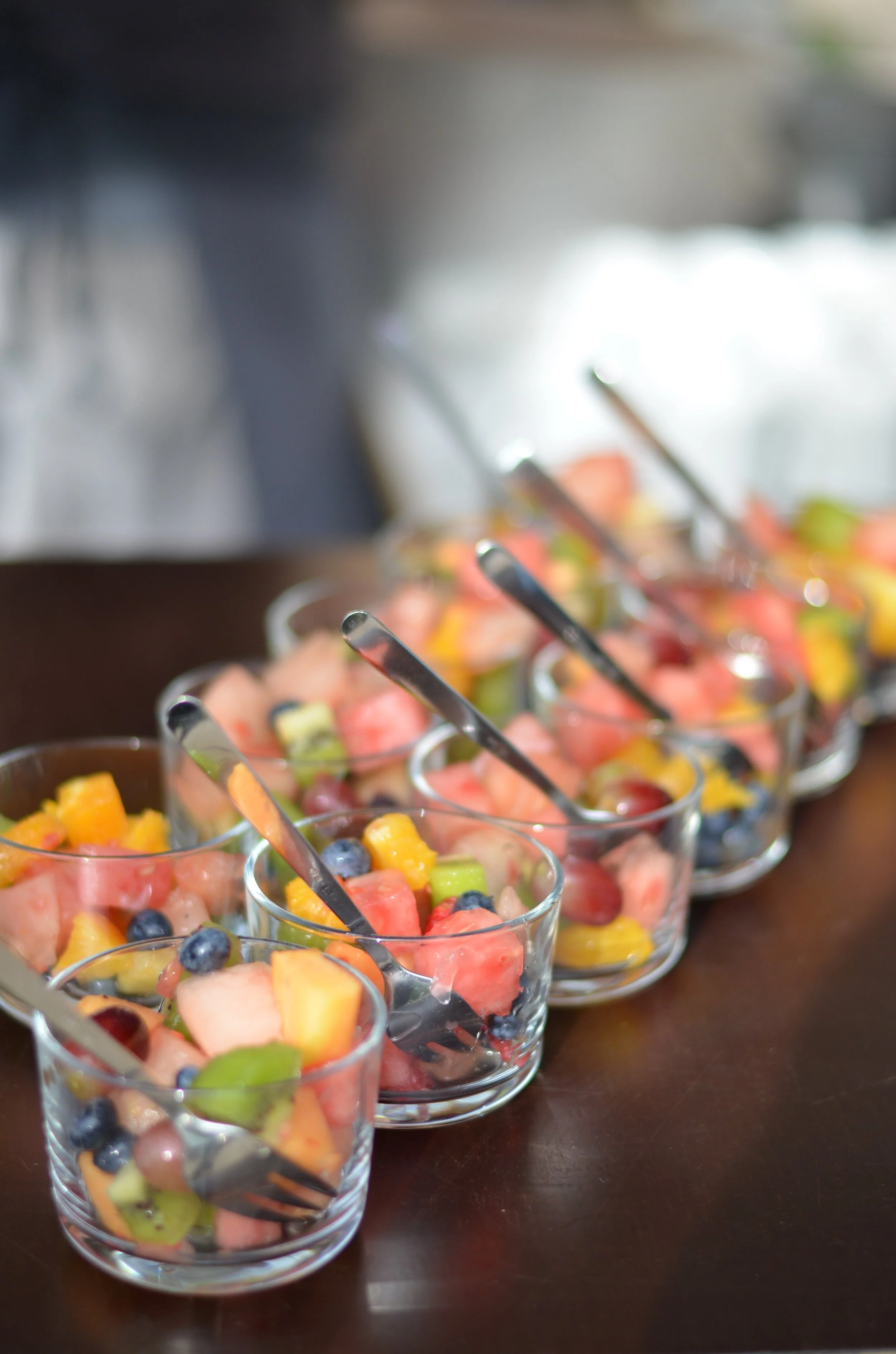 Mehrere Gläser mit Obstsalat, gefüllt mit Kiwi, Melone, Trauben, Mango und anderen Früchten, auf einem dunklen Tisch