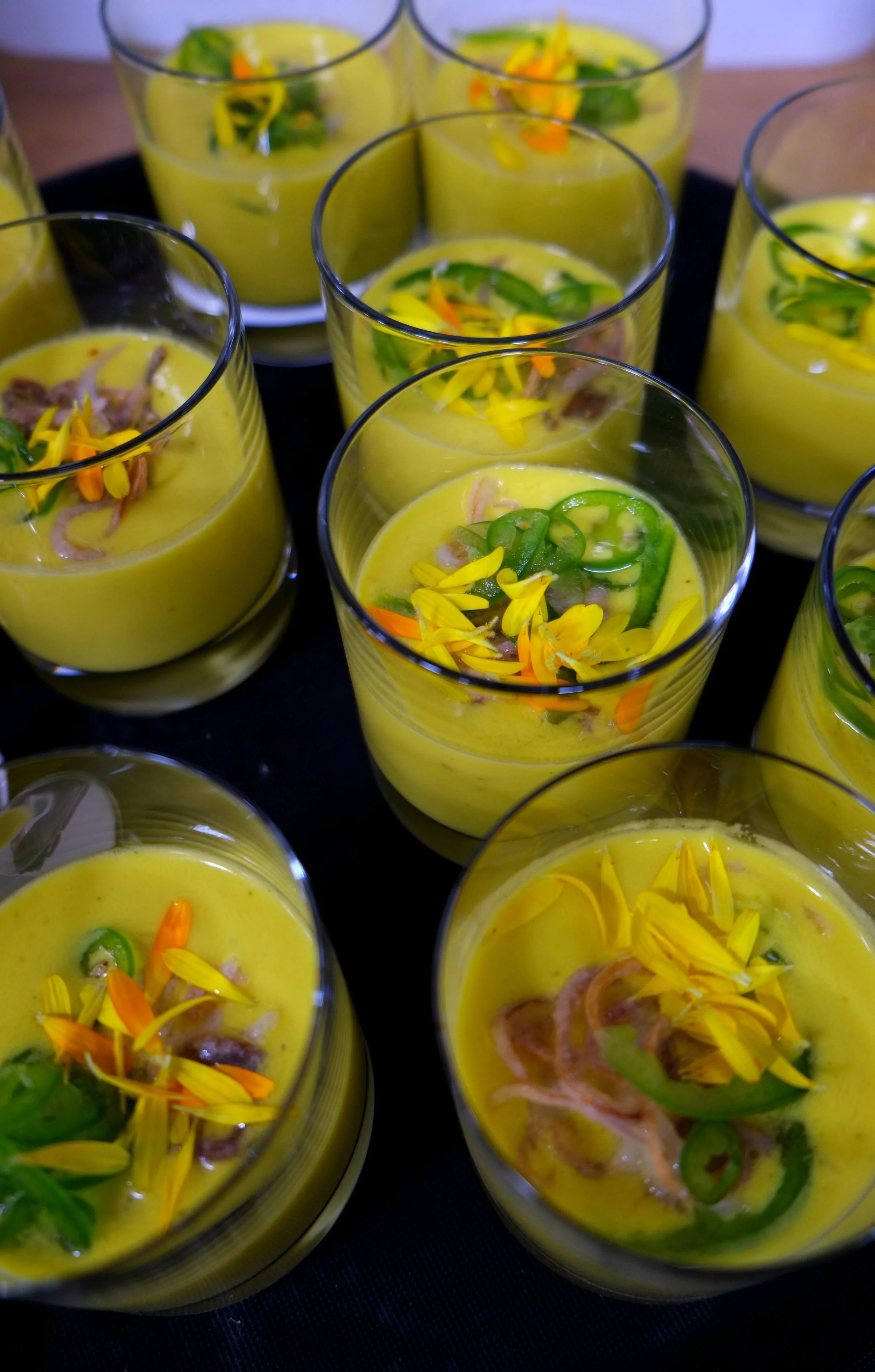 Mehrere Gläser mit einer gelben Suppe, garniert mit grünen und orangefarbenen Blumen und grünen Chili-Schoten.