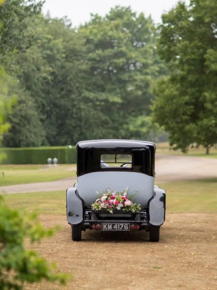 Vintage black and gray car with a flower arrangement on the back, parked on a dirt road surrounded by greenery.