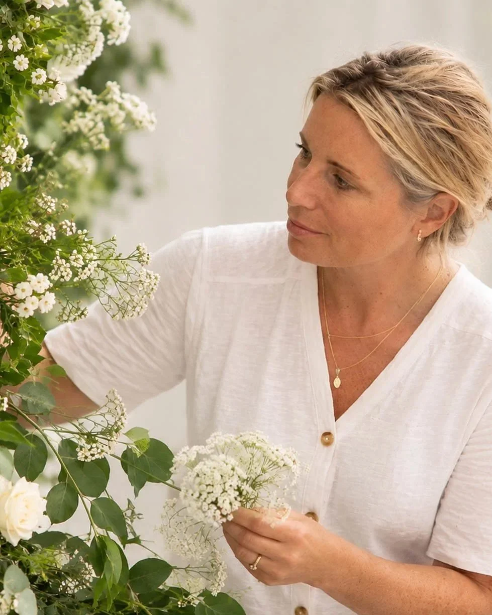 A woman with short, blonde hair wearing a white top and gold jewelry gently arranging white flowers among green foliage.