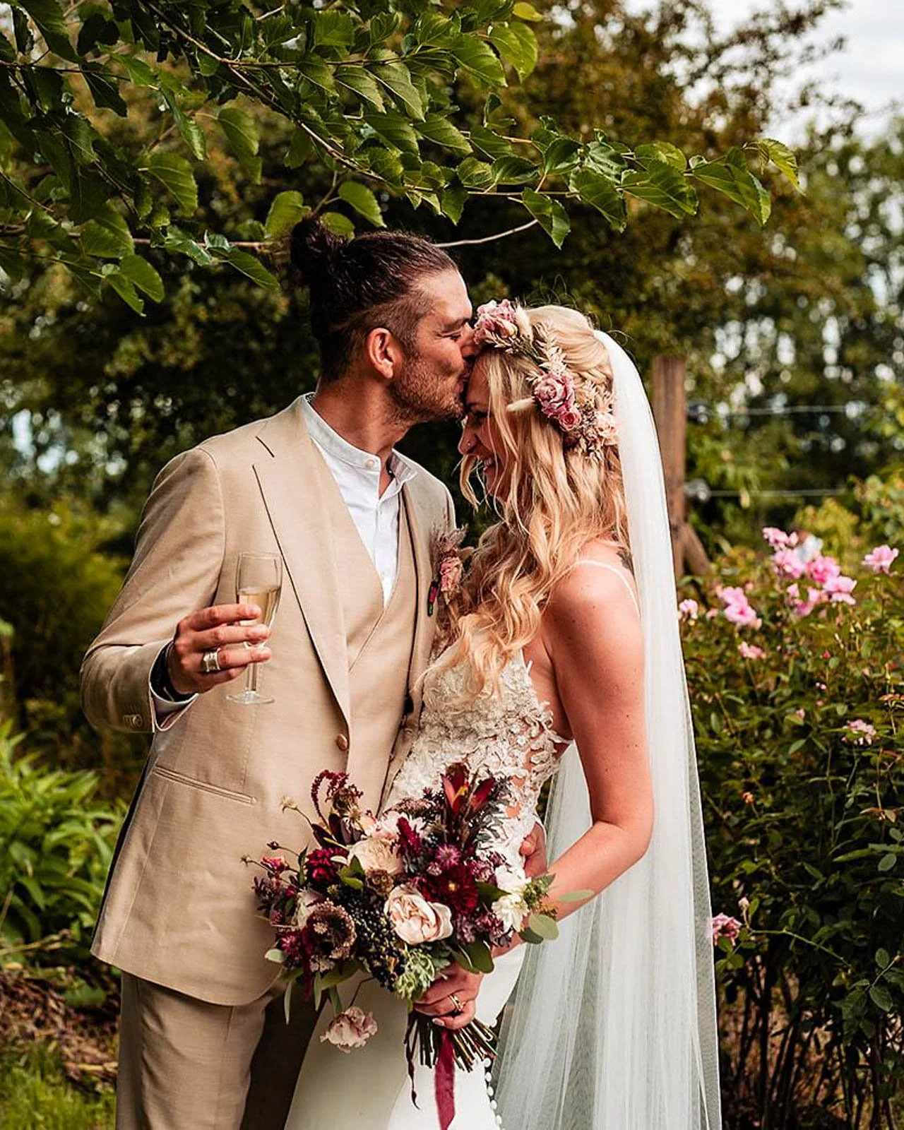 A bride and groom sharing a kiss outdoors, with the groom wearing a beige suit and the bride in a lace wedding dress, holding a bouquet of flowers, surrounded by greenery and pink flowers.