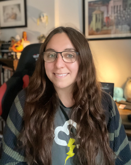 Woman with long brown wavy hair, glasses, and a grey T-shirt with a graphic, sitting in a home office with a gaming chair, shelves, and artwork in the background.