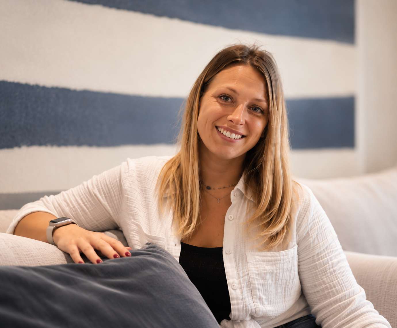 Une femme souriante avec des cheveux longs, assise sur un canapé beige dans un intérieur moderne.