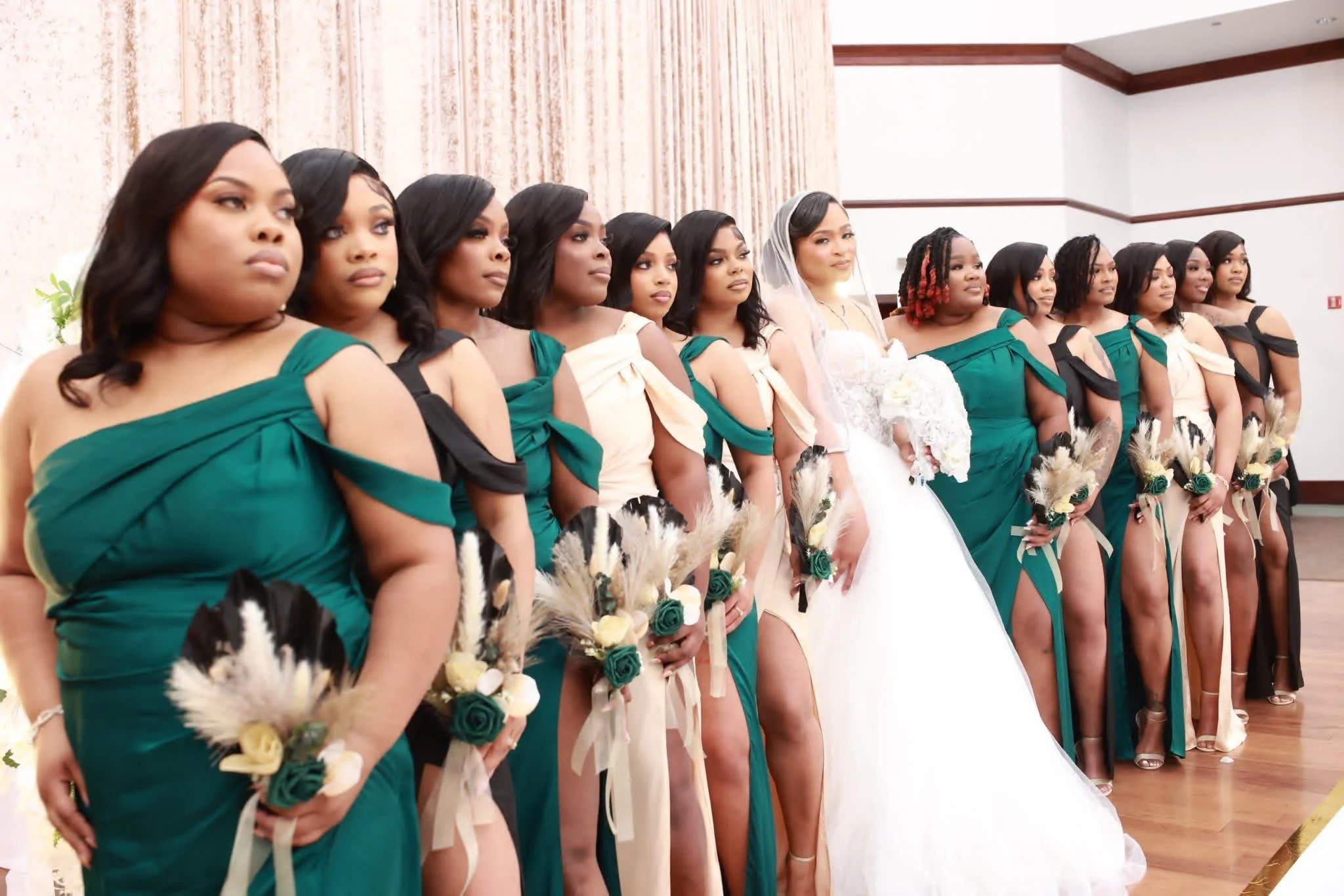 A bride in a wedding dress and veil stands among bridesmaids, all holding bouquets, in a reception hall.