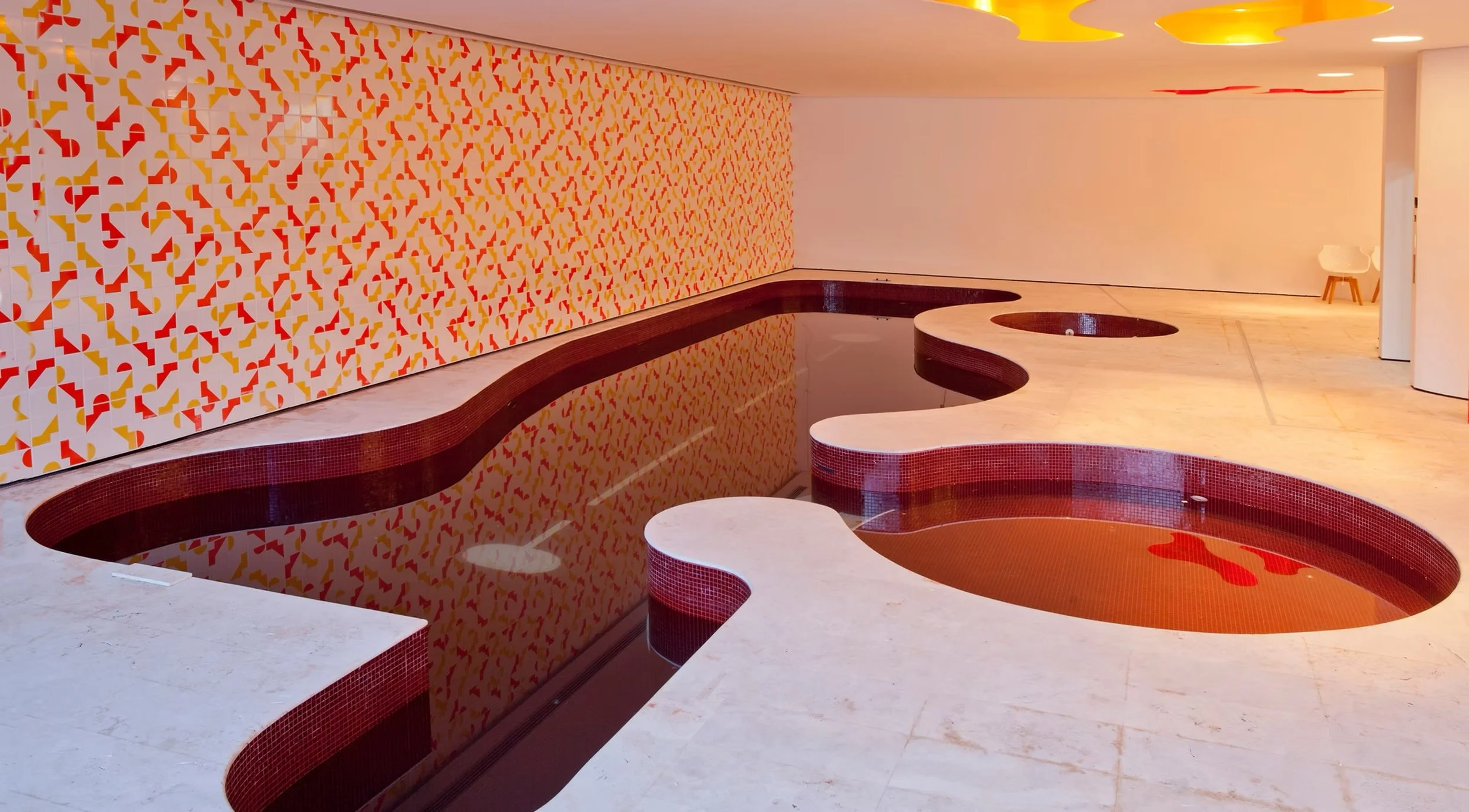 Piscina com formato irregular revestida com pequenas azulejos vermelhos, ao lado uma parede de mosaico colorido em tons de vermelho, amarelo e branco, iluminação de teto e cadeira branca ao fundo.