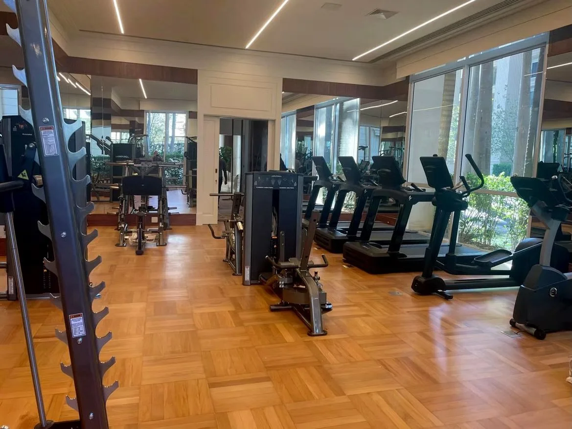 Sala de ginásio com equipamentos de cardio, como esteiras e bicicletas ergométricas, com janelas grandes e piso de madeira.