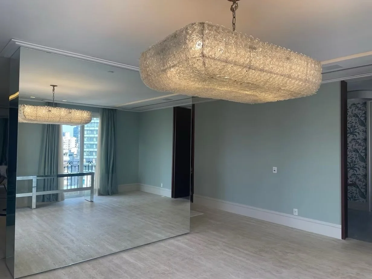 Salão de apartamento com lustres de cristal, paredes verdes claras, piso de madeira clara, espelho grande na parede e varandinha com vista da cidade.