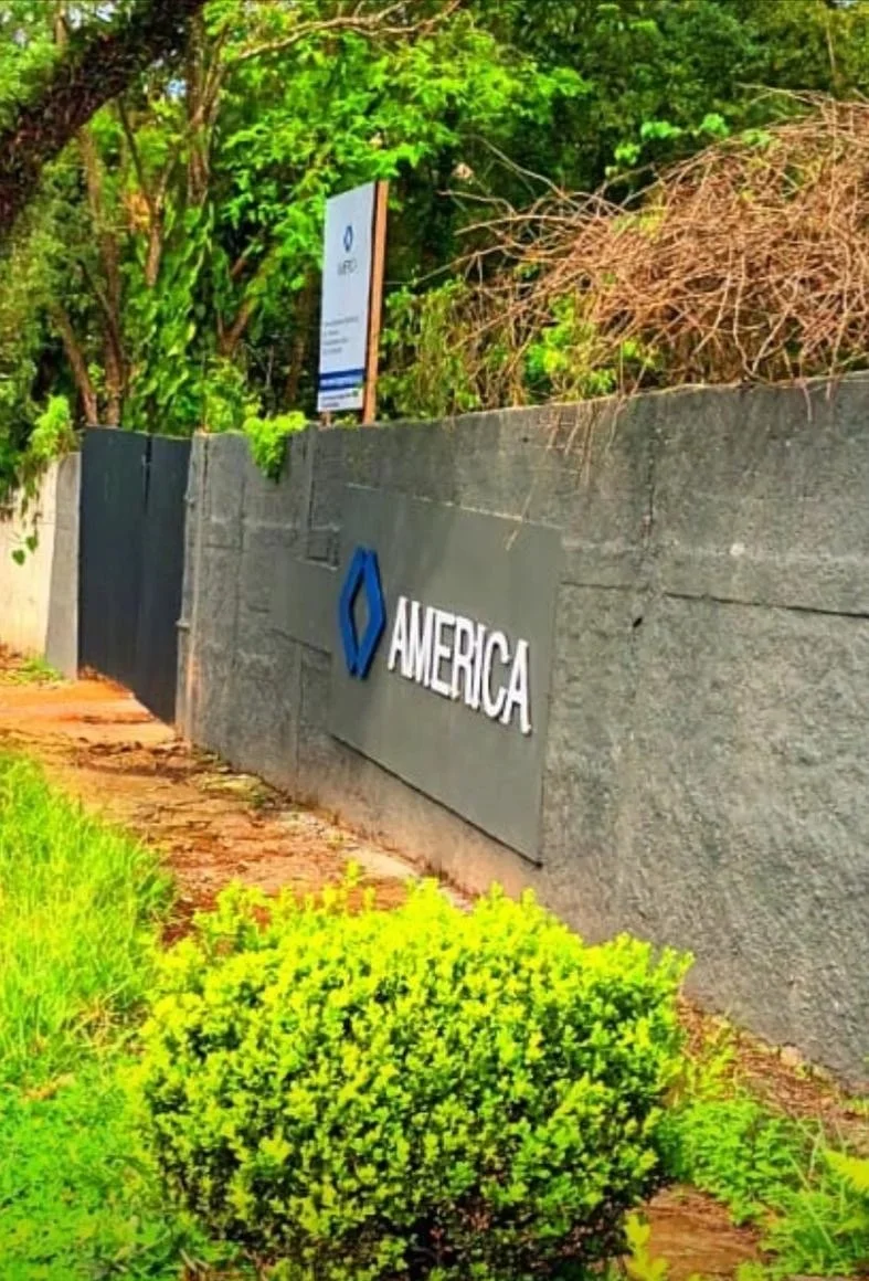 Muro de concreto com placa de sinalização e logo da empresa Amerca, com vegetação ao redor e árvores ao fundo.