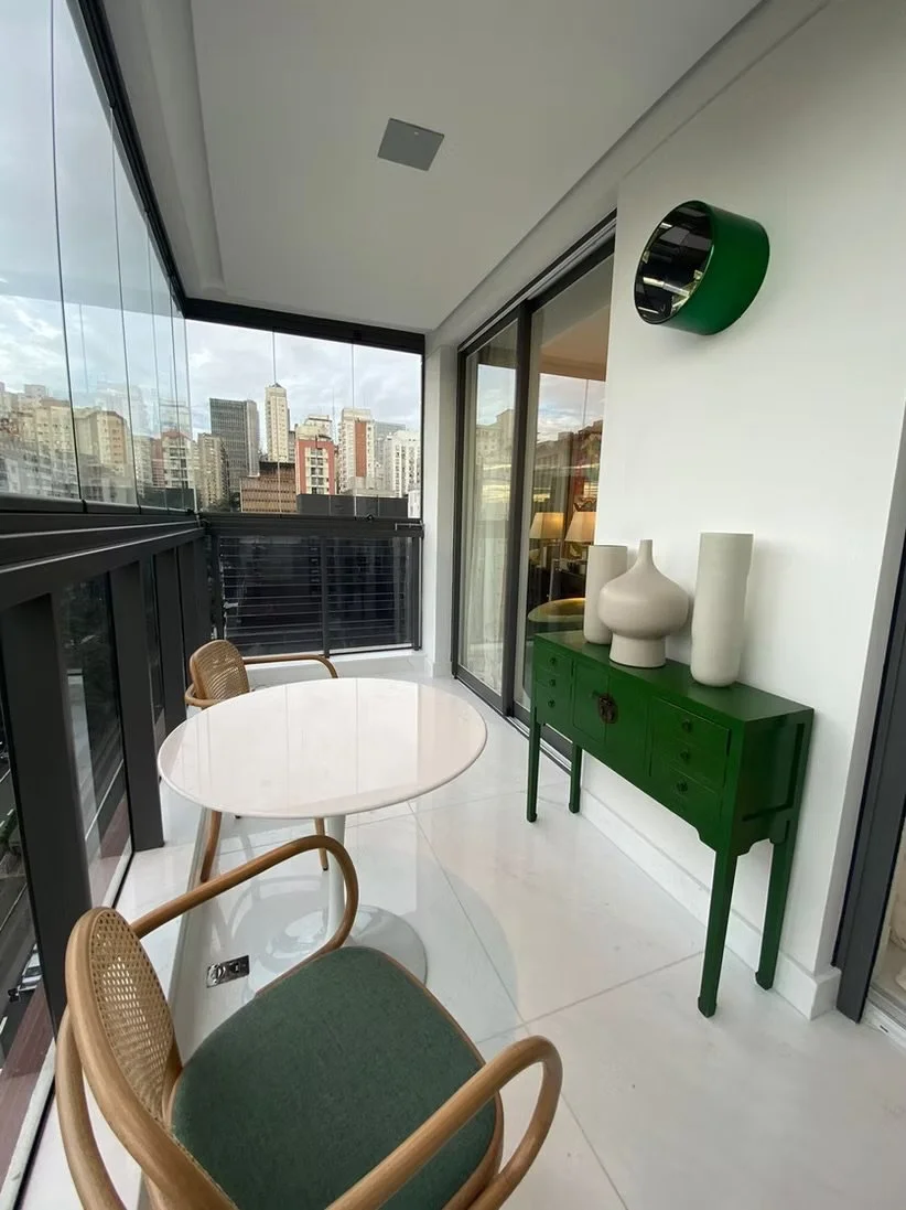 Varanda com mesa de vidro redonda, duas cadeiras de madeira com assentos verdes, armário verde e vasos decorativos brancos, vista de prédios urbanos ao fundo.