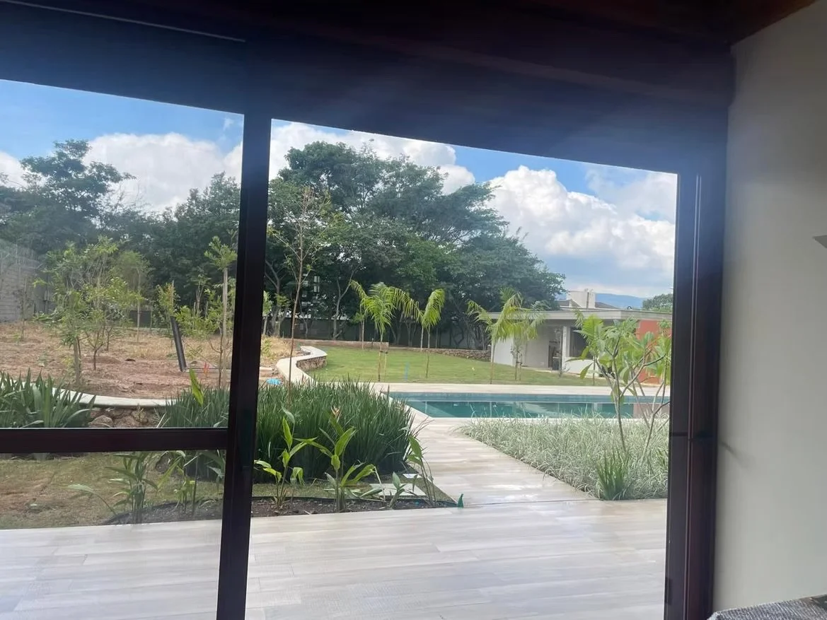 Vista de um jardim com piscina vista de uma janela de vidro de uma casa, árvores e céu com nuvens.