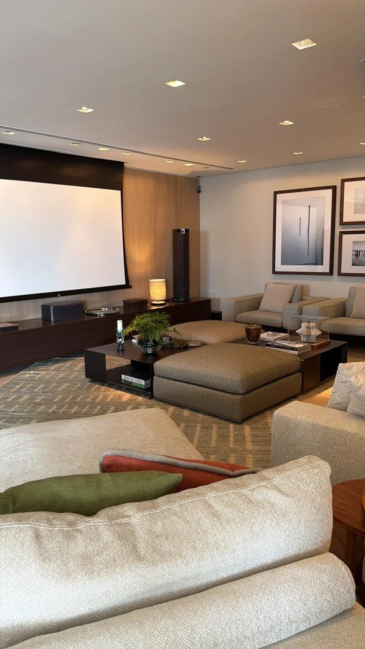 Sala de estar moderna com sofá, poltronas, uma mesa de centro decorada com plantas, livros e objetos decorativos, além de uma tela de projeção na parede e quadros na parede ao fundo.
