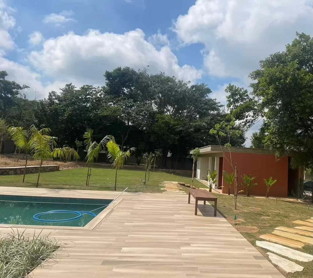 Área de lazer com piscina, cadeiras de madeira, jardim com árvores e plantas, estrutura de construção ao fundo, céu com nuvens.