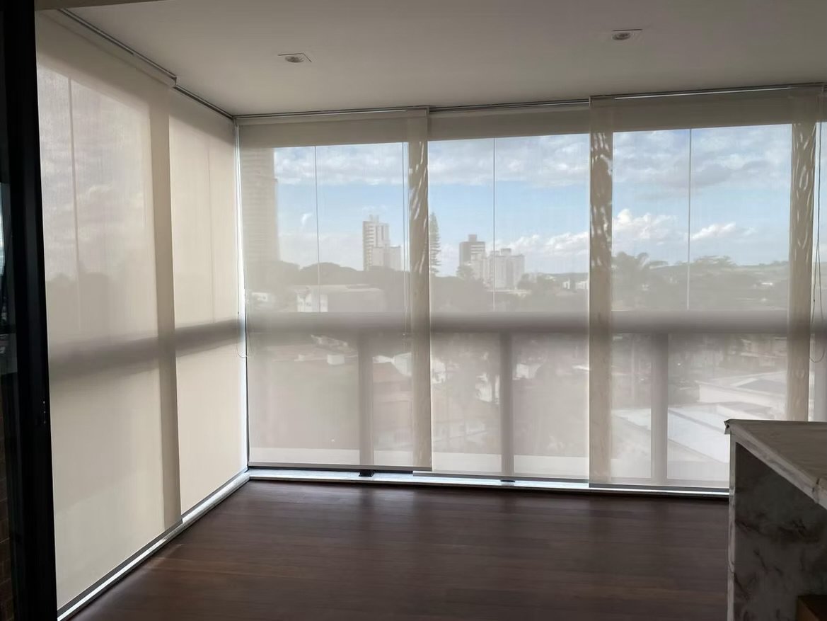 Varanda de apartamento com cortinas transparentes, piso de madeira escura e vista para uma cidade com prédios e céu ensolarado.