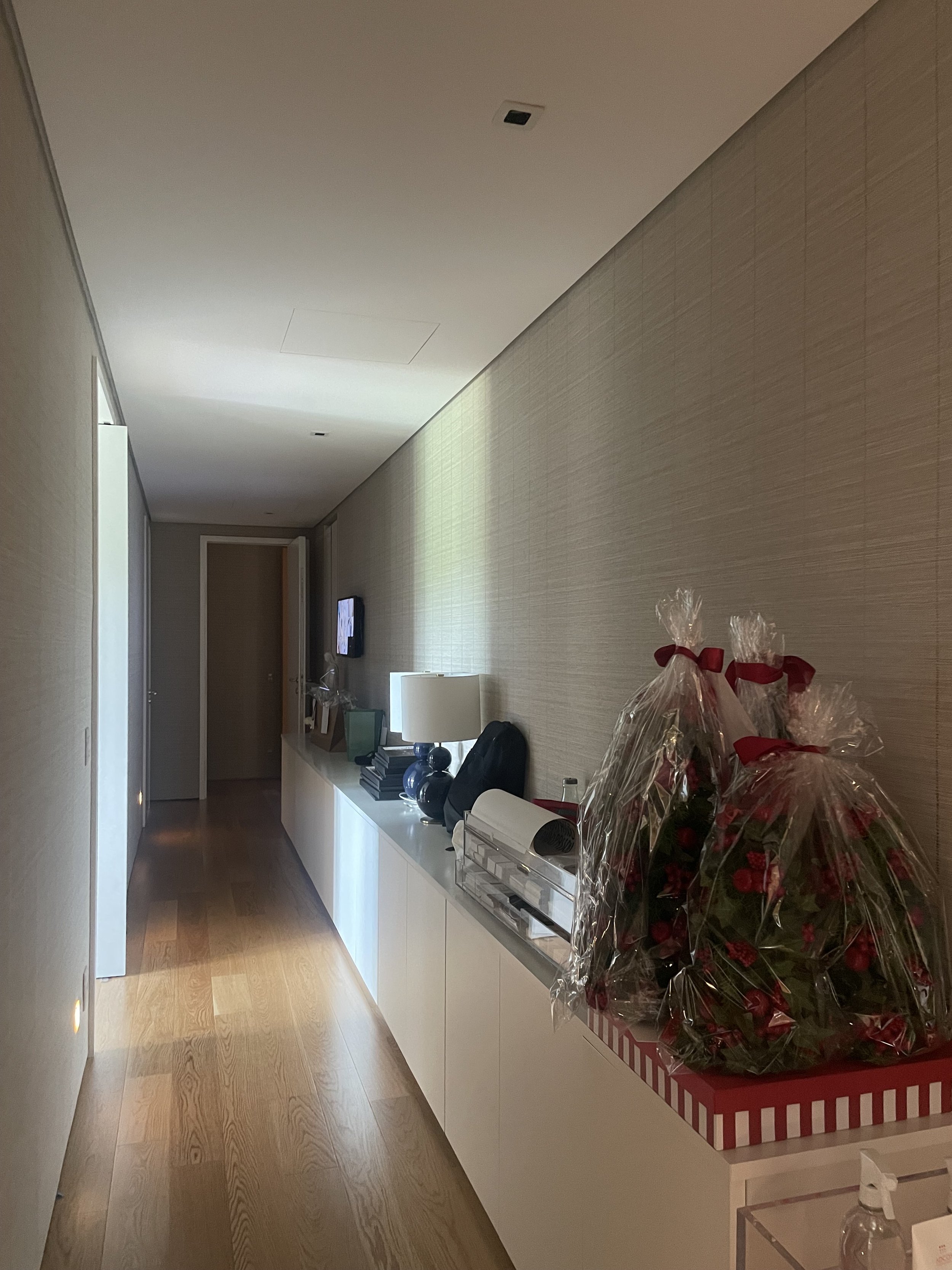 Corredor de um apartamento com piso de madeira, paredes claras, uma televisão presa na parede do fundo, uma mesa branca ao lado direito com uma luminária e uma caixa de presentes com embalagem de plástico e fitas vermelhas.